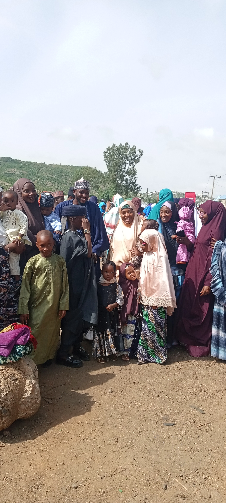In Naraguta Hausawa, Nigeria, a group of people is gathered outdoors. The group includes men, women, and children of varying ages, dressed in traditional Muslim attire. The women mostly wear hijabs of different colors, including brown, peach, and purple. The men wear caps and robes, some with glasses. Some people hold babies or young children. The ground is dirt. Behind the group are hills and a few buildings. The sky is overcast.