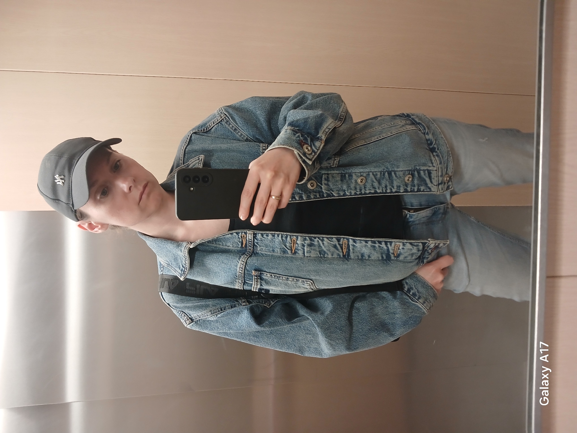 A person in a gray baseball cap, blue denim jacket, and blue jeans is taking a selfie in a mirror. The person is holding a black smartphone. The reflection reveals an elevator-like interior with a panelled wall on one side and a mirrored surface on the other. The phone displays a "Galaxy A17" inscription in the lower right corner of the reflection. The image lacks sufficient contextual information to determine if the location is Yekaterinburg, Russia.
