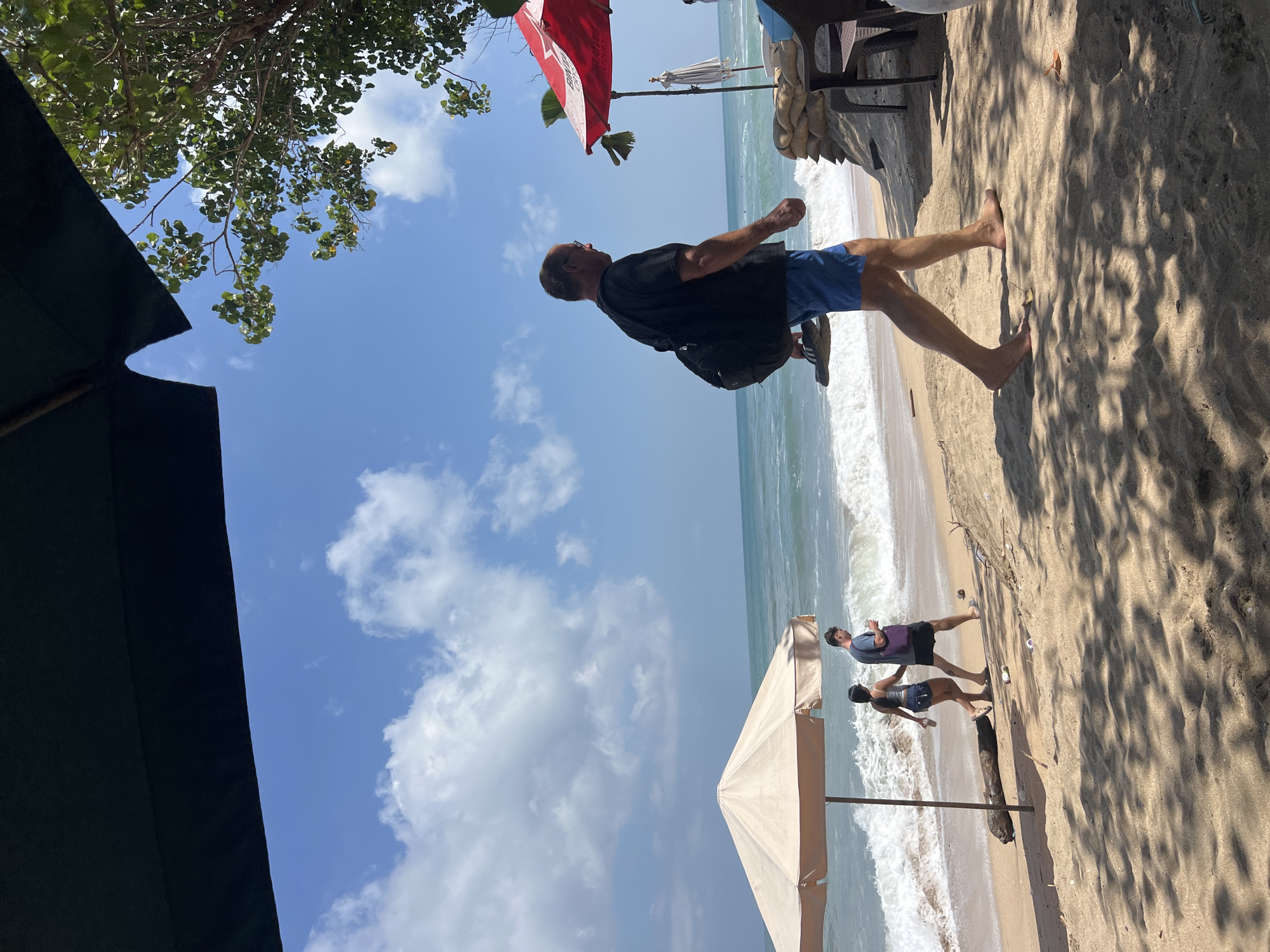 In Kuta, Indonesia, on a sunny day, the scene depicts a beach with people walking along the shore. A man in a black t-shirt and blue shorts walks towards the right, carrying a black backpack over his left shoulder and a pair of flip-flops in his right hand. Further down the beach, a couple walks towards the left, with the woman in a white tank top and denim shorts, and the man in a gray t-shirt and dark shorts, carrying a purple bag.
Two beach umbrellas provide shade on the sandy shore. A large, light-colored umbrella is to the left of the frame, with the couple walking beneath it. A red umbrella, partly visible on the right, is positioned near a table and chairs, suggesting a seating area. The ocean with gentle waves extends to the horizon under a blue sky with scattered clouds. The foreground is shaded by foliage, casting dappled shadows on the sand. The overall atmosphere is relaxed, indicating a typical beach scene in Kuta.