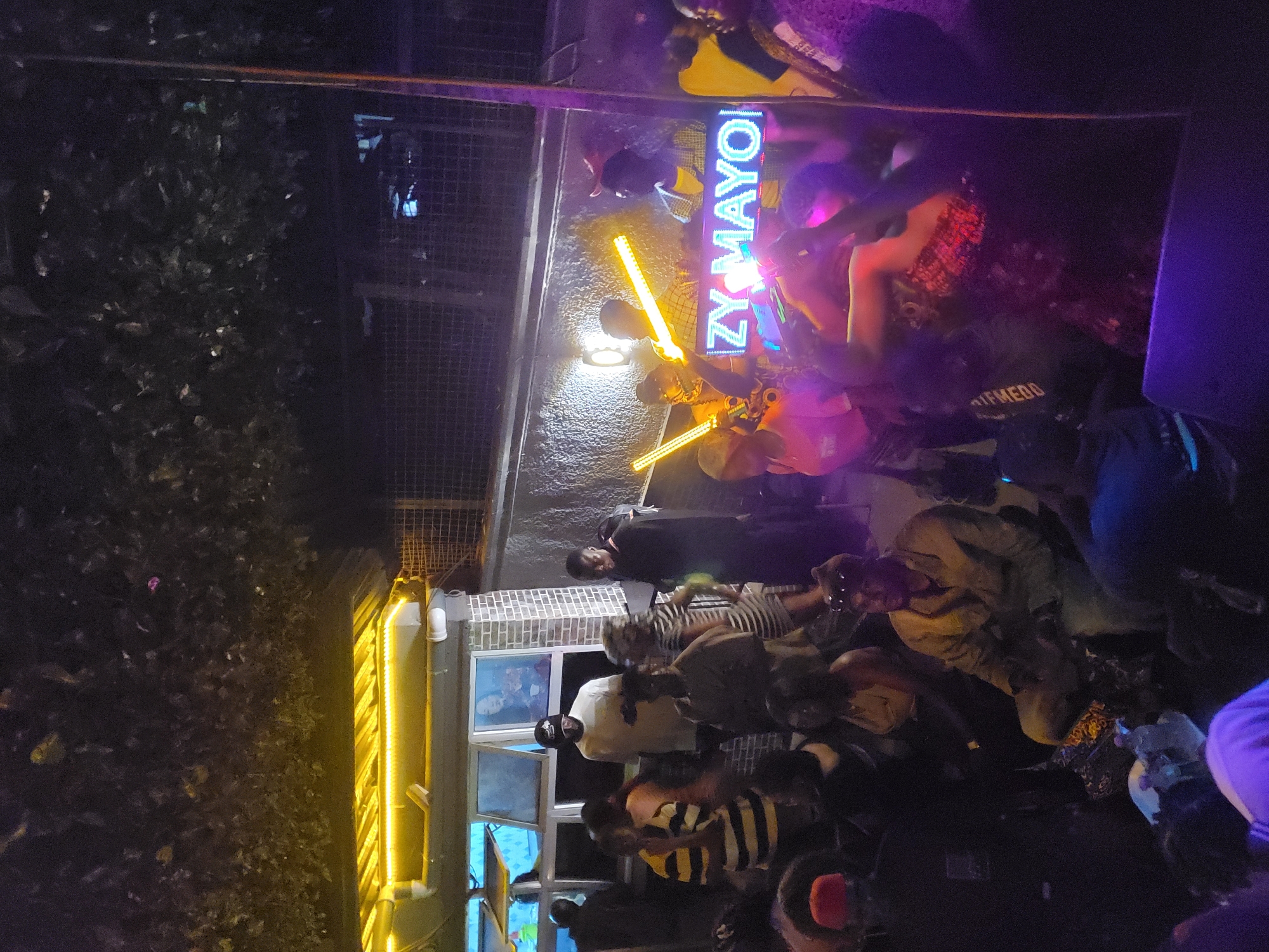 A nighttime view in Nnewi, Nigeria, shows a gathering of people outside "ZY-MAYO," an establishment identified by a neon sign. People are standing and congregating in front of a building with windows displaying blue light inside. A bright yellow light strip illuminates the entrance. Trees are visible on the left side. The individuals in the foreground appear to be in casual attire. Two people are holding light sticks.