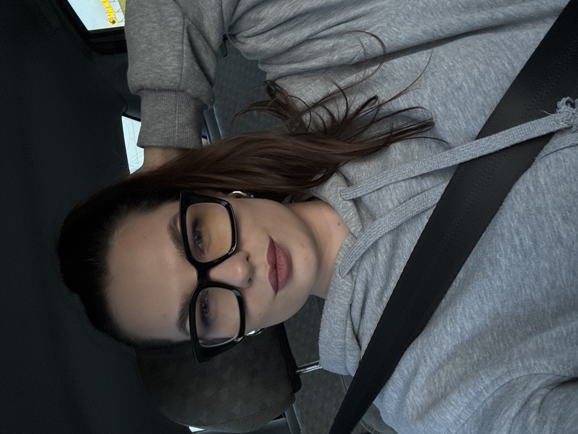 A woman with long brown hair pulled into a ponytail, wearing oversized black-framed glasses and a grey hooded sweatshirt, is pictured from the chest up. She is inside a vehicle, evidenced by a seatbelt across her torso and the car's interior visible in the background, including a headrest and a window. Her left arm is raised, resting her head, and she has subtle makeup on her lips. The lighting suggests it is daytime, possibly late afternoon or early evening, with a soft glow coming from the window. The environment appears to be a typical urban street setting, with a blurry yellow sign visible through the window, hinting at a commercial establishment. There are no other people or animals visible in the image. The scene is a selfie, taken by the woman herself.
