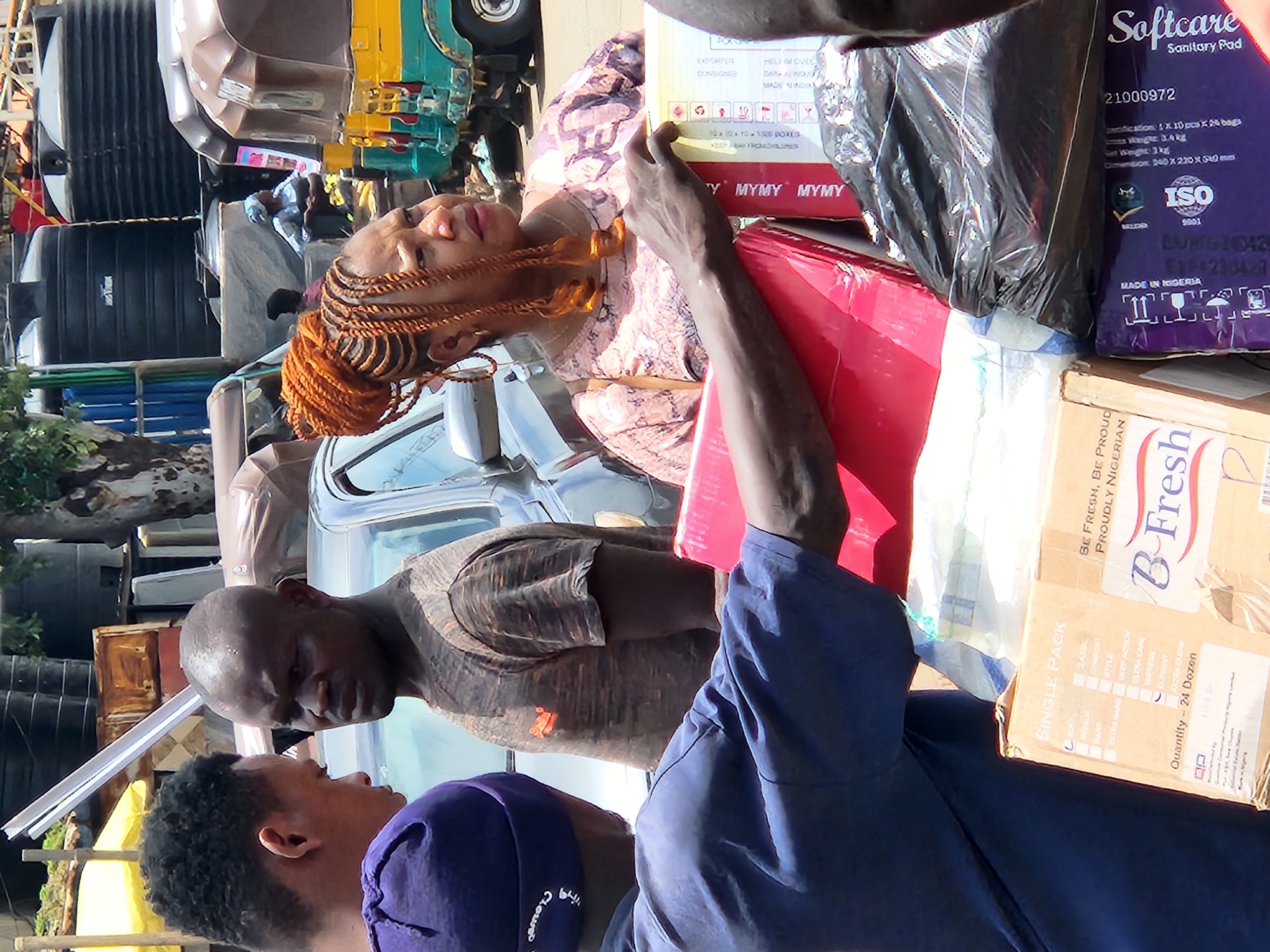 This is a close-up, eye-level shot of three people and various items on a street in Jos, Nigeria. The foreground is dominated by the back of a person wearing a blue shirt and a purple beanie, carrying a red box. To their right, a person in a pink and purple patterned shirt with elaborate braided hair is also holding a box. In the mid-ground, a man with a shaved head and wearing a grey t-shirt faces towards the left, appearing to interact with the person in the blue shirt. Behind them, a car and several commercial vehicles, including a yellow and green auto-rickshaw, are visible. In the background, large black water tanks and other structures suggest an urban setting. The overall lighting indicates daytime.