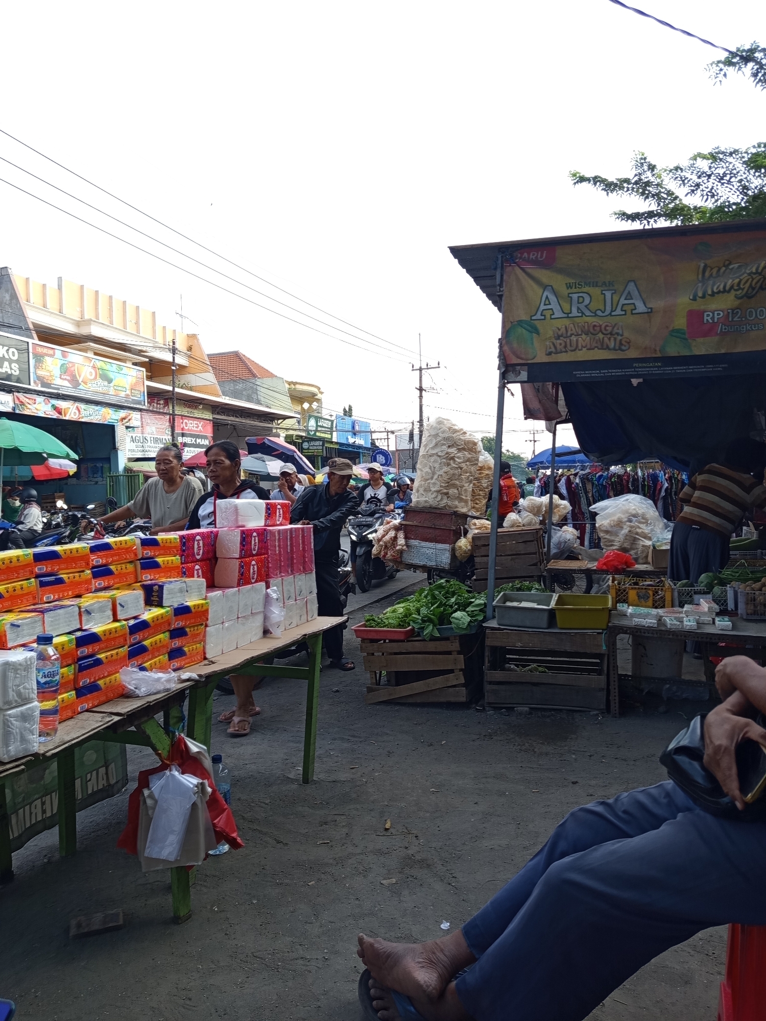 The image depicts a bustling street market in Suko, Indonesia. In the foreground, a table laden with stacks of tissue paper products is prominently displayed. A man in a dark jacket and cap is seen pushing a cart towards the right side of the frame, while another man in a light-colored shirt stands behind the tissue paper table, seemingly engaged in a transaction. Behind them, a variety of market stalls are visible, offering goods such as fresh produce, snacks, and clothing. Several motorcycles are parked or being ridden through the market. The environment appears to be an open-air setting with buildings and awnings lining the street. The overall scene conveys a lively and active atmosphere typical of a local Indonesian market.