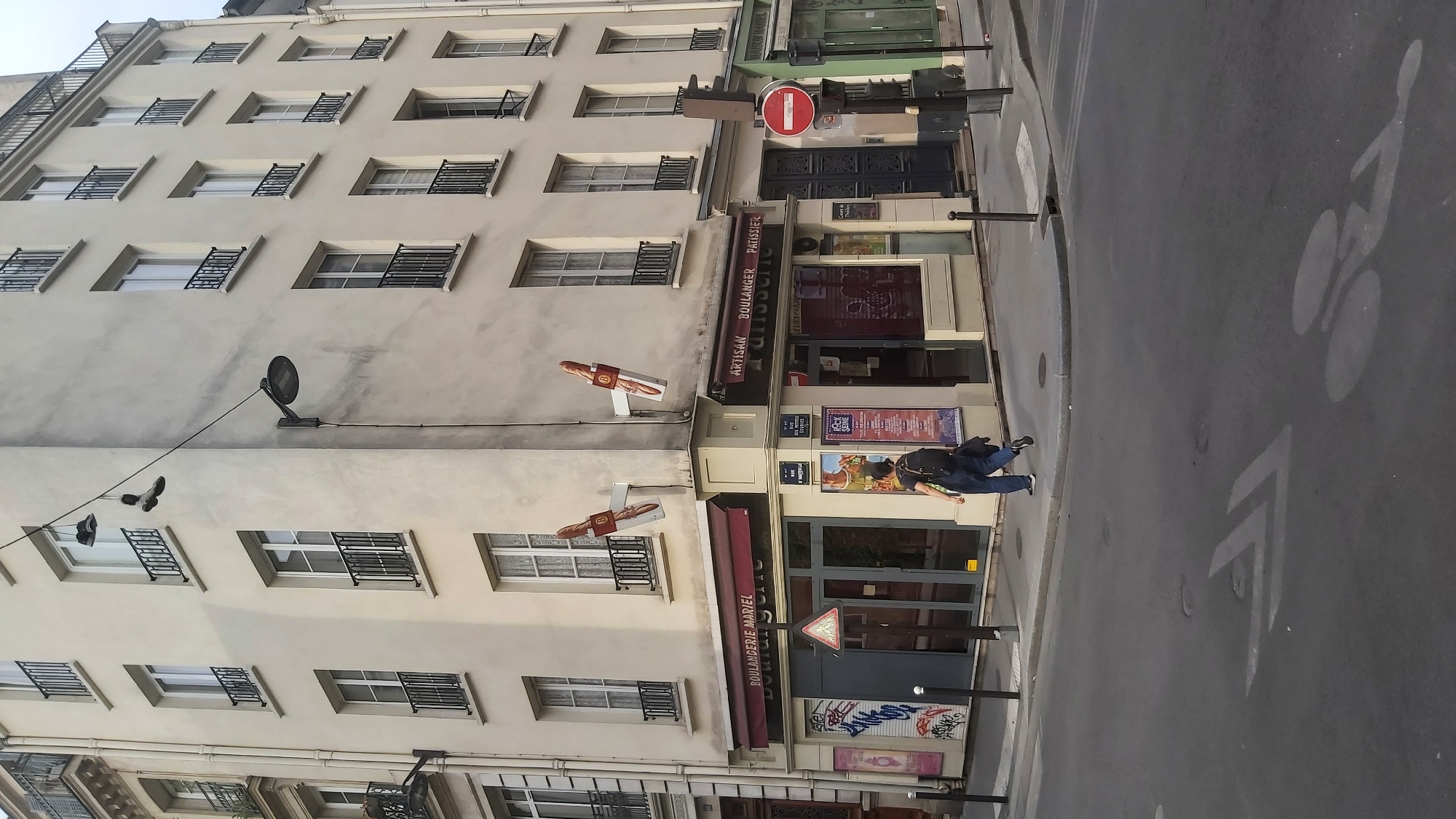 This is a medium shot, eye-level, outdoor, daytime, medium-angle photograph of a street corner in Paris, France. The scene is dominated by the cream-colored facade of a multi-story building that occupies the corner of an intersection. The building has multiple floors with evenly spaced windows, each featuring black wrought-iron railings. On the second floor, a street lamp is mounted on the corner of the building, and a pair of old shoes are suspended from a wire extending from this lamp across the side of the building.
At street level, the building houses two adjacent businesses: a "BOULANGERIE MARIEL" on the left and an "ARTISAN BOULANGER PATISSERIE" on the right. Both businesses have awnings and glass storefronts. The "BOULANGERIE MARIEL" storefront features graffiti on the left side and a triangular warning sign in front. The "ARTISAN BOULANGER PATISSERIE" storefront displays posters and has a red and white circular "no entry" sign visible on a pole to its right. Two baguette-shaped signs are mounted on the exterior of the building above the storefronts, pointing outwards.
A lone individual, viewed from behind, walks away from the camera and towards the right side of the frame, crossing the street. The person is wearing dark pants, a dark backpack, and dark shoes. The street itself is asphalt, with painted lane markings and a bicycle lane symbol visible. The overall impression is a typical Parisian street scene with a mix of residential and commercial elements.
