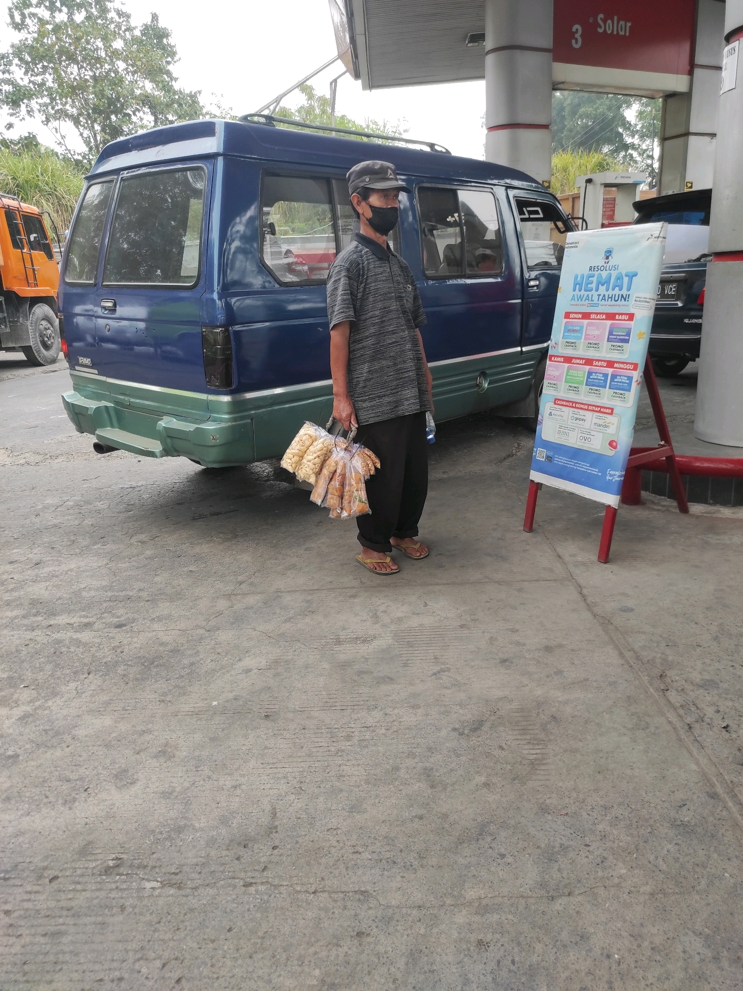 A man wearing a black face mask, a grey striped t-shirt, black pants, a dark baseball cap, and yellow sandals stands in front of a blue van at a gas station. He is holding a bunch of bagged snacks in his right hand. A sign advertising promotions is to his right. The ground is paved concrete. The background shows trees and a portion of the gas station canopy with a "3 Solar" sign. An orange truck is partially visible to the left of the blue van. The scene appears to be located in Padalarang, Indonesia, at a Pertamina gas station, indicated by the signage and the context of the region.