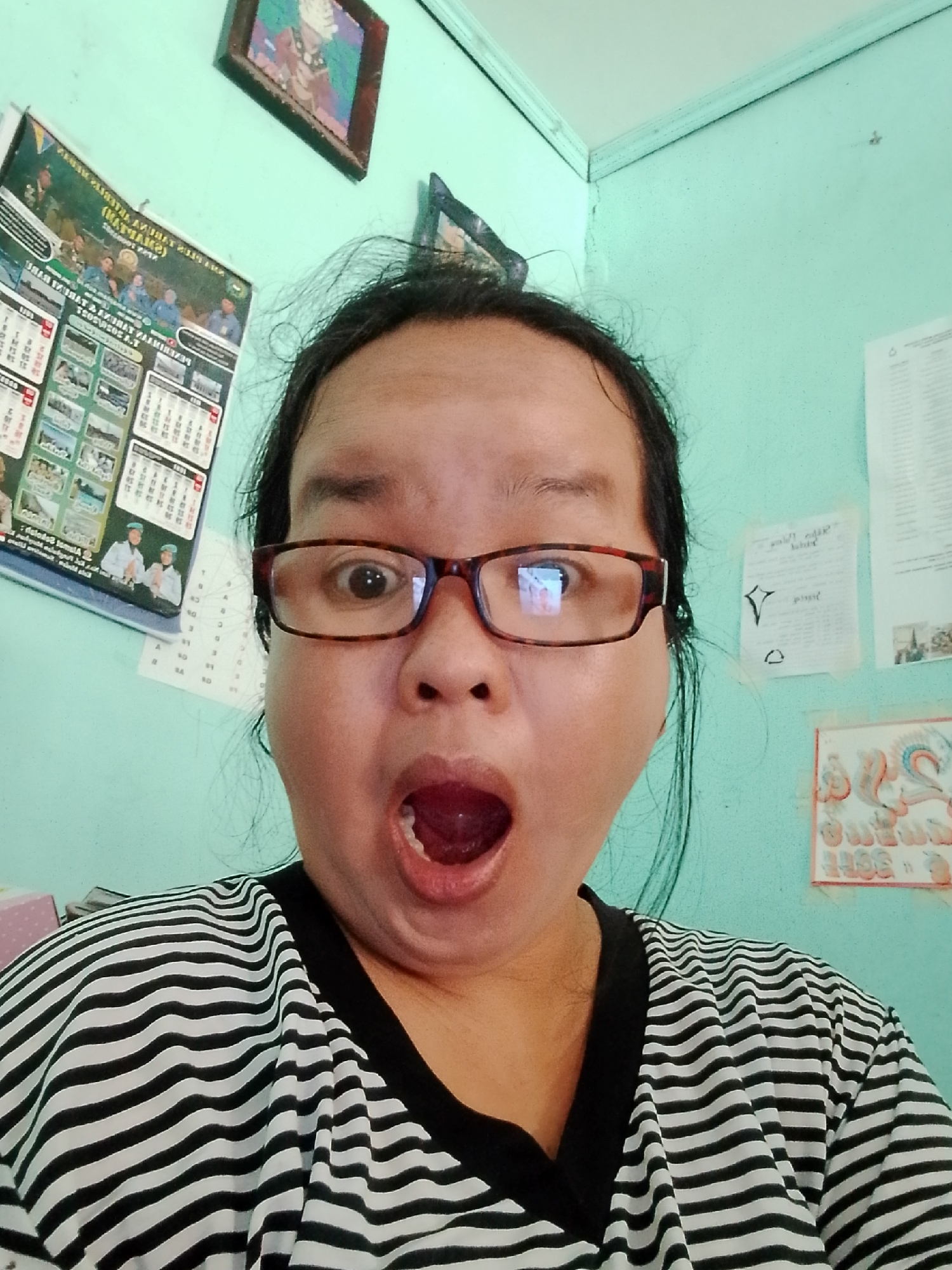 A young woman with black hair and glasses makes a surprised face at the camera, her mouth open in an 'O' shape. She is wearing a black and white striped t-shirt. The background is a light teal wall with various papers and a framed picture hanging on it. To the left of the woman, a calendar with pictures and text is attached to the wall. The scene appears to be indoors, possibly in a home or office, in the city of Serbelawan, Indonesia.