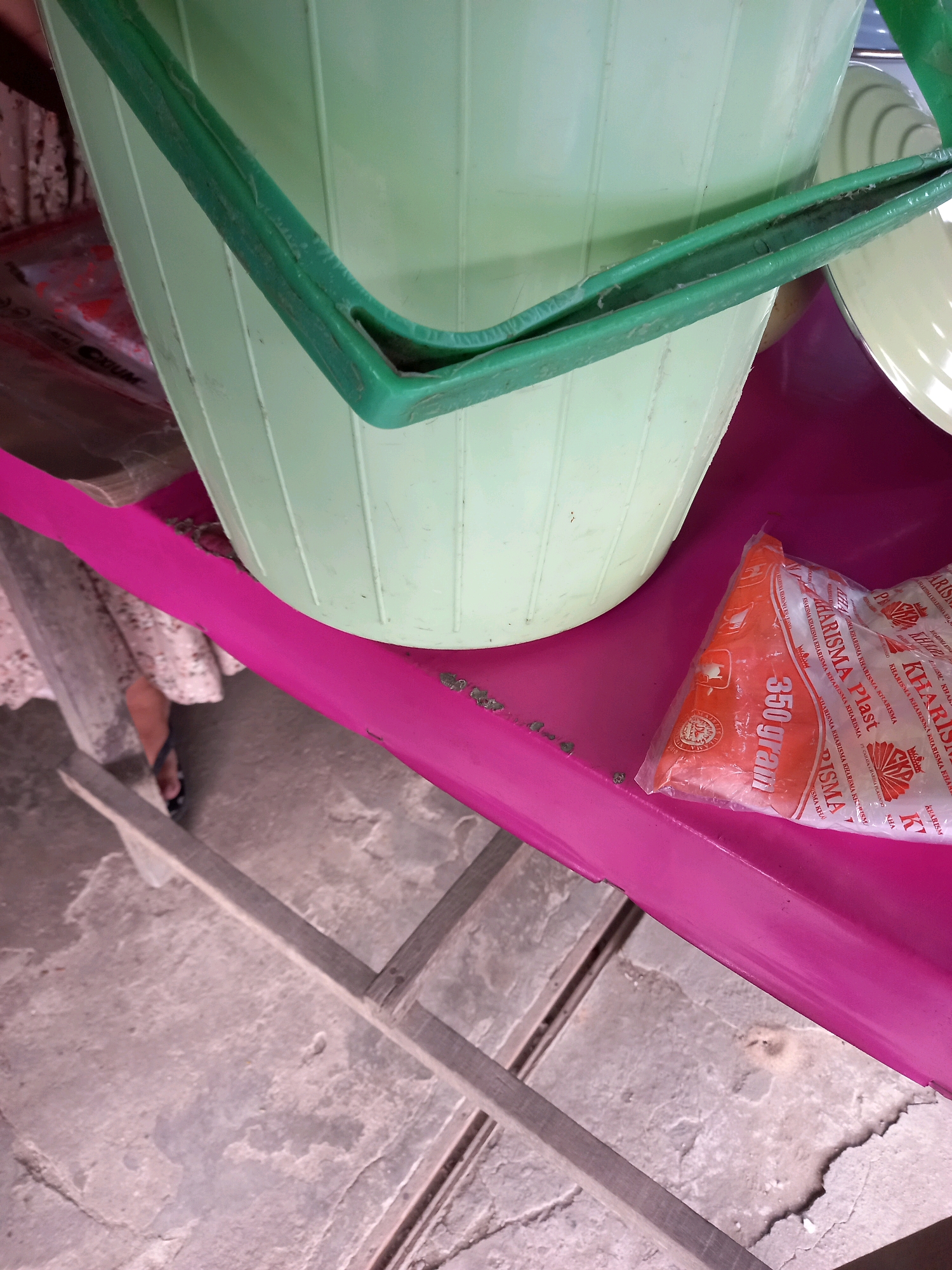 A light green bucket with a green handle rests on a vibrant pink surface, which appears to be a part of a mobile cart or stall. The bucket is positioned to the left of the frame, partially obscuring a white and yellow patterned bowl on its right. In the foreground, to the right of the bucket, is a clear plastic package containing an orange product. The packaging displays red text with the brand name "KHARISMA Plast" and indicates "350 gram."
The pink surface extends across the lower half of the image, with a wooden structure visible beneath it, suggesting a market stall or outdoor display. The ground appears to be a rough, concrete surface, possibly a sidewalk or pavement. A person's legs, wearing a light-colored floral dress and black flip-flops, are visible on the far left, partially behind the bucket. The scene is brightly lit, suggesting it is daytime. The overall setting implies a commercial environment, likely a market or shop in the city of Samarinda, Indonesia, given the context provided.