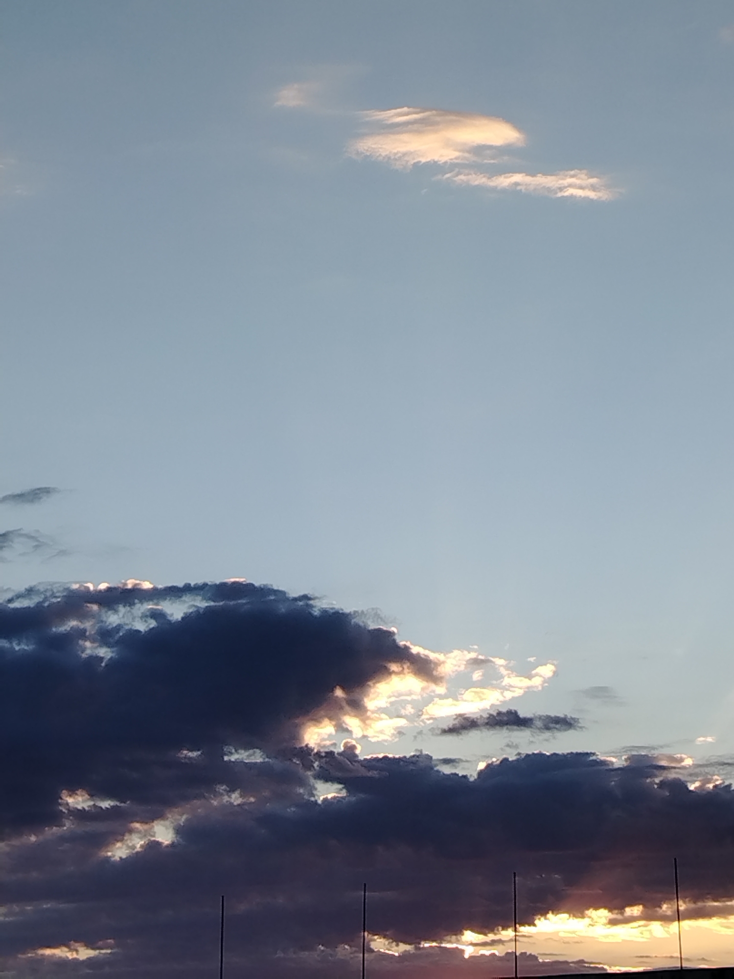The image shows a sky with clouds at sunset over Baikonur, Kazakhstan. The clouds are dark and dramatic, with some areas illuminated by the setting sun. The sky is a pale blue, transitioning to hues of orange and yellow near the horizon. There are also some lighter, wispy clouds higher up. The foreground is dark and indistinct, with the tops of what appear to be poles visible. The scene evokes a sense of natural beauty and the vastness of the sky.