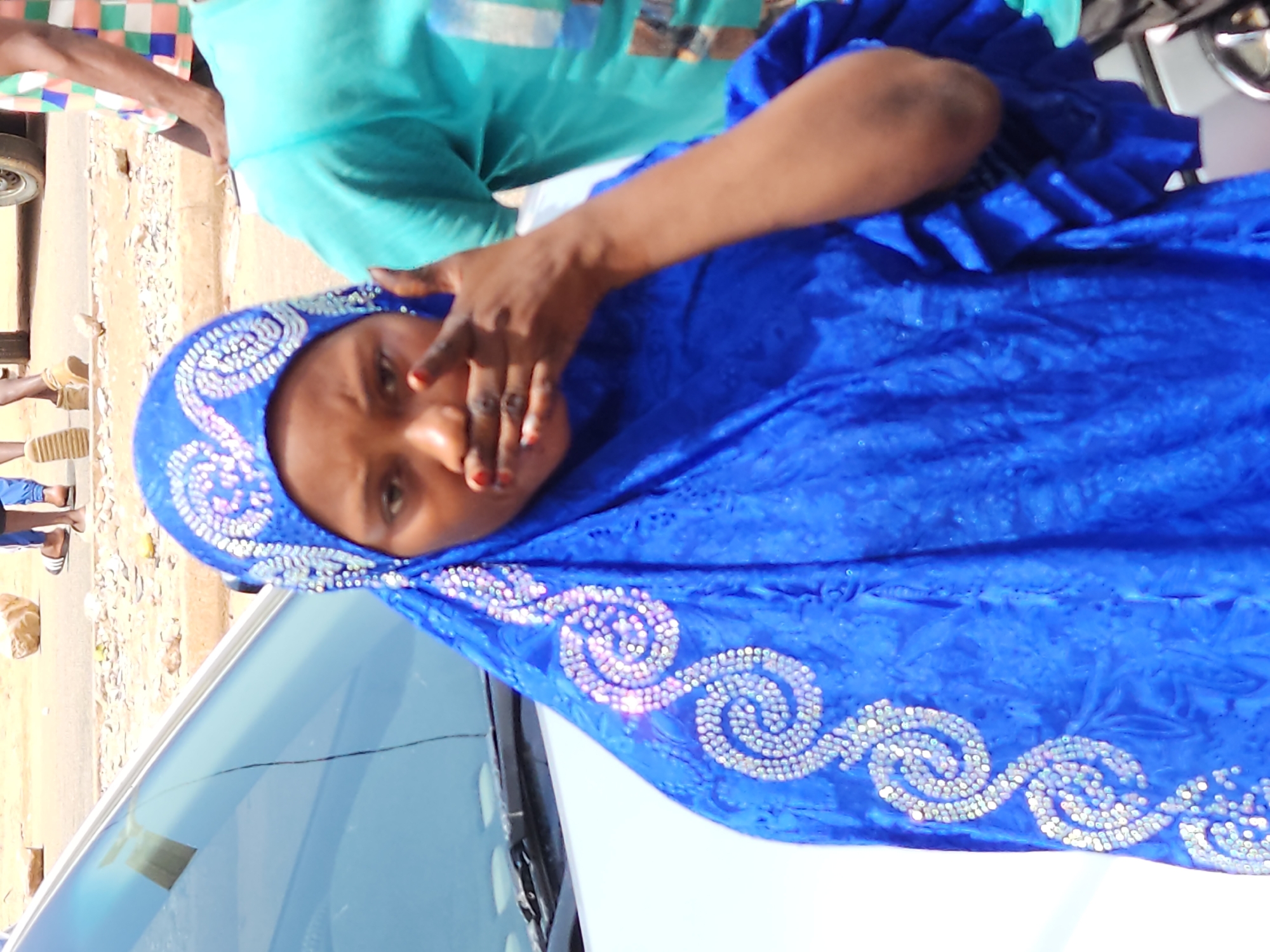 A woman, possibly of African descent, is looking at the camera, covering her mouth with her right hand, suggesting surprise or shock. She is wearing a vibrant blue headscarf and dress, both adorned with intricate silver embellishments. The headscarf is a hijab, covering her hair and neck, and the dress appears to be a loose-fitting garment. The woman stands in front of a white car, its windshield visible. In the background, blurred figures and a street scene suggest an outdoor urban environment. The overall impression is one of a candid, somewhat dramatic moment captured in an everyday setting.