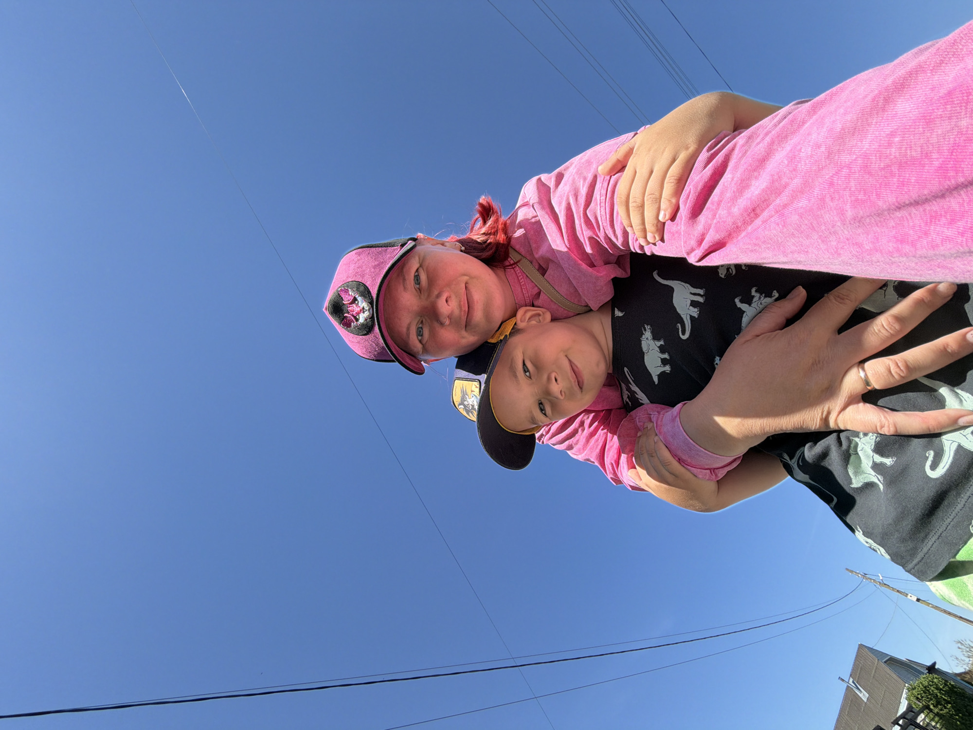 A low-angle shot depicts a woman and a child, likely a mother and her son, embracing. The woman wears a pink cap with a graphic on the front and a matching pink top. Her red hair is visible under the cap. The boy wears a dark baseball cap with a golden emblem and a black t-shirt with dinosaur prints. They are both facing the camera, smiling slightly.
The background is a clear blue sky intersected by electrical power lines. In the lower part of the frame, the corner of a building with a roof and a bit of green vegetation can be seen. The scene appears to be outdoors, possibly in a residential area of Chaikovsky, Russia.