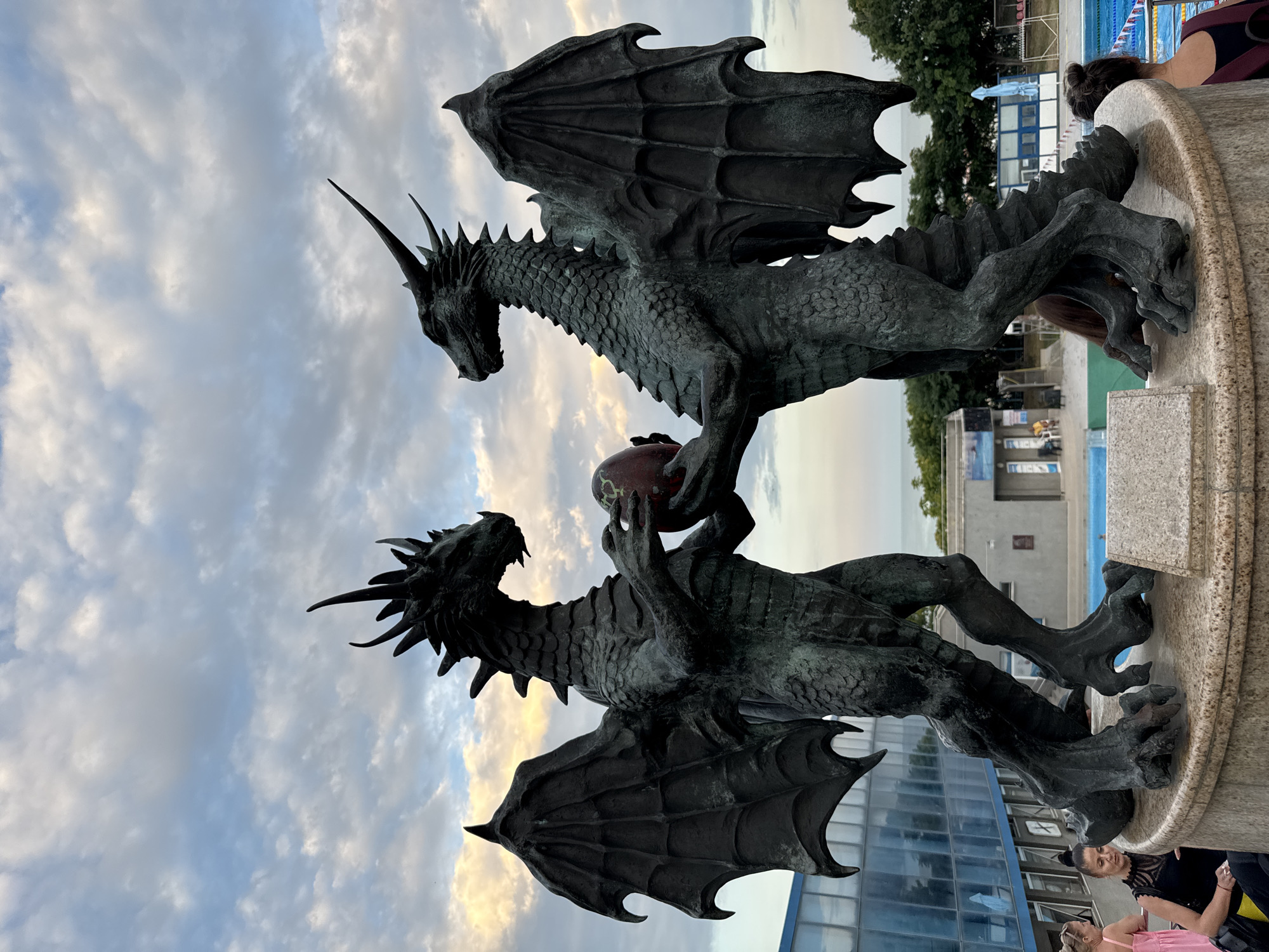 This image captures a striking bronze sculpture of two dragons, positioned as if in a struggle or interaction, with one dragon appearing to hold a reddish orb. The sculpture is placed outdoors, with a cloudy sky visible in the background, suggesting it is either morning or late afternoon. In the distance, a blue swimming pool and some buildings indicate a public or recreational setting, possibly a park or a complex with aquatic facilities. The detailed scales and formidable wings of the dragons are prominent features.
Several people are visible in the foreground and background, some appearing to be spectators or passersby. Their clothing and hairstyles suggest a casual, everyday atmosphere. One woman in the foreground, with her hair tied up, is looking towards the sculpture.
The overall mood is one of awe and perhaps a touch of mystery, evoked by the dramatic and powerful depiction of the mythical creatures. The light from the setting or rising sun casts a warm glow on the clouds. There is no visible text in the image. The location context is Varna, Bulgaria.