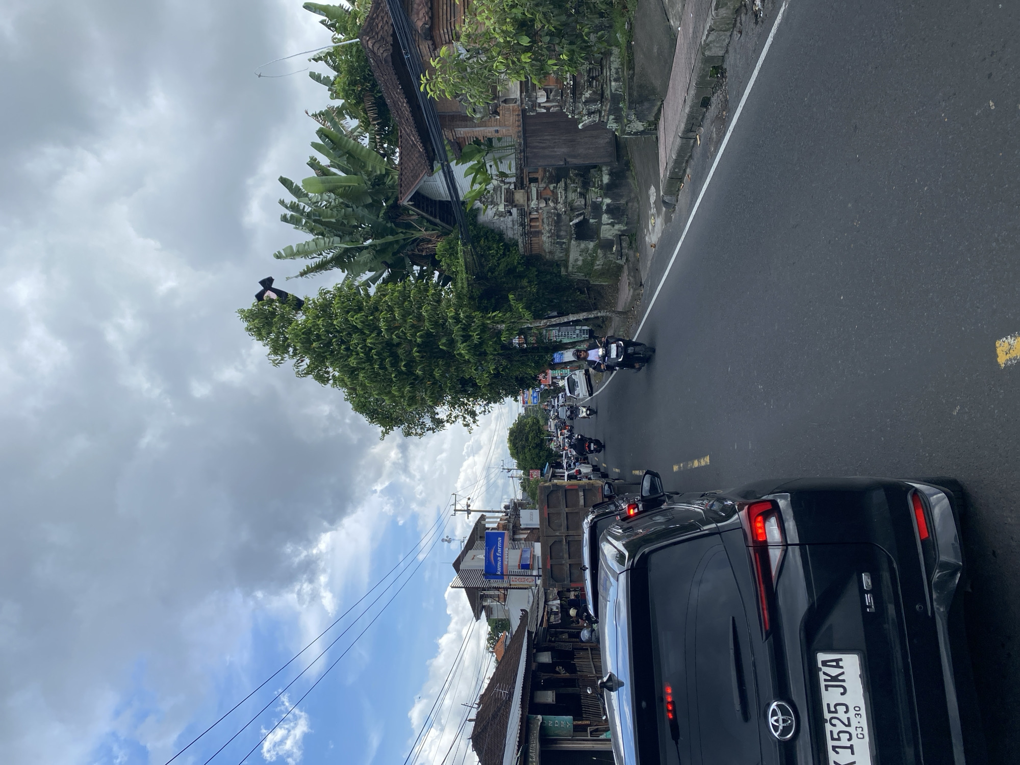 The image depicts a street scene in Ubud District, Indonesia. A black car is prominently featured in the foreground with its license plate, "K 1525 JKA," visible. The car is stationary in what appears to be traffic. Other vehicles, including scooters and cars, are visible further down the road, suggesting congestion. Buildings typical of Balinese architecture are on either side of the road, with some showing commercial signage. Overhead power lines are strung along the road. The sky is partly cloudy. People are seen on the scooters and as pedestrians. The weather seems overcast. Trees and tropical foliage are interspersed among the buildings. The road is asphalt.