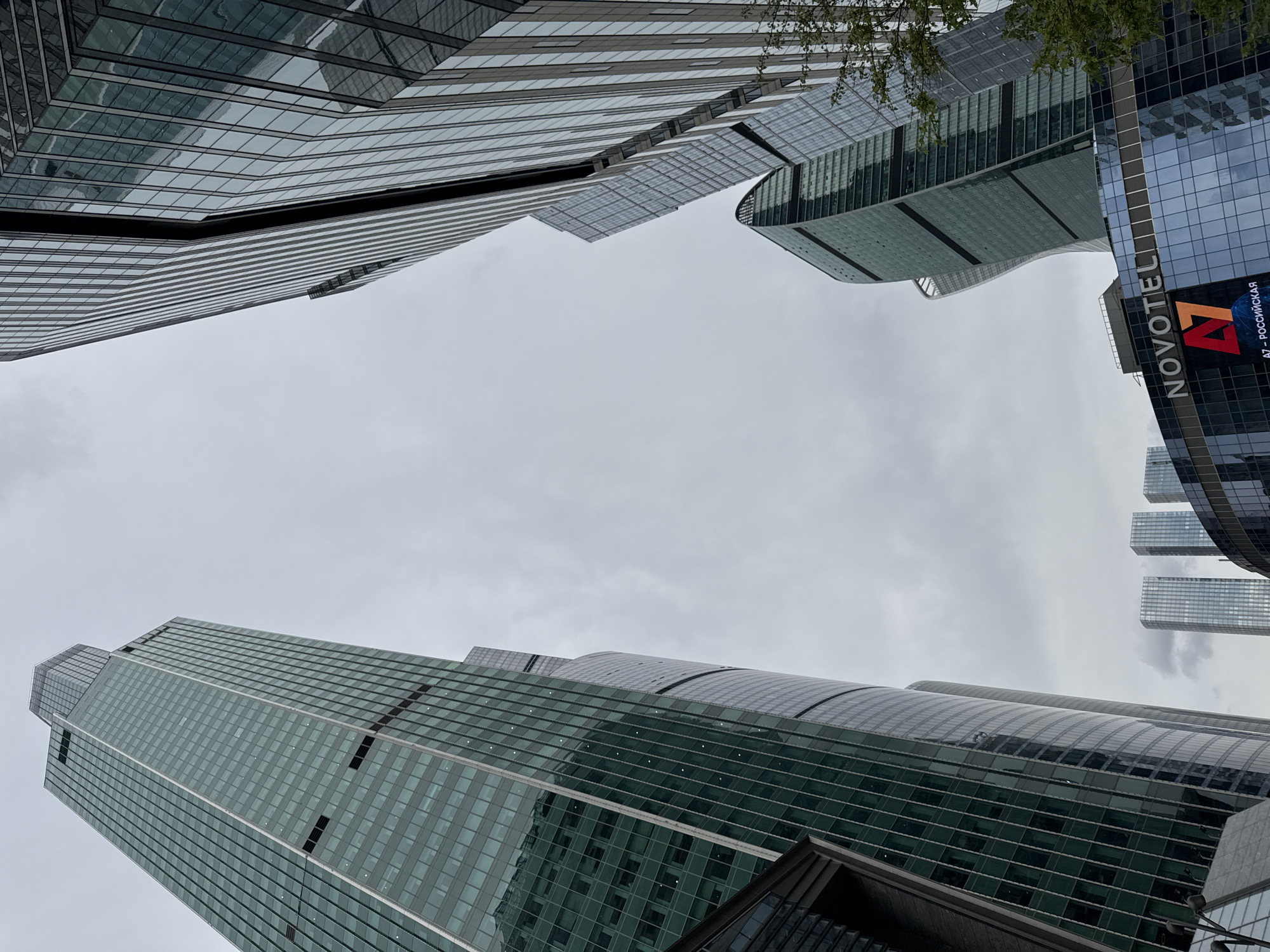 The image is a low-angle shot of several skyscrapers in a city, possibly Moscow, Russia, as indicated by the Novotel А7 - РОССИЙСКАЯ sign on one of the buildings. The buildings are modern and mostly glass-clad, reflecting the overcast sky. The composition emphasizes the height and density of the buildings, which angle towards the center of the frame, creating a converging perspective. The sky is a flat gray color, suggesting a cloudy day. The presence of the Novotel sign, along with the modern architectural style, suggests this is a commercial or business district. There is no presence of people in this image. There is no clear indication that this is in Khimki.