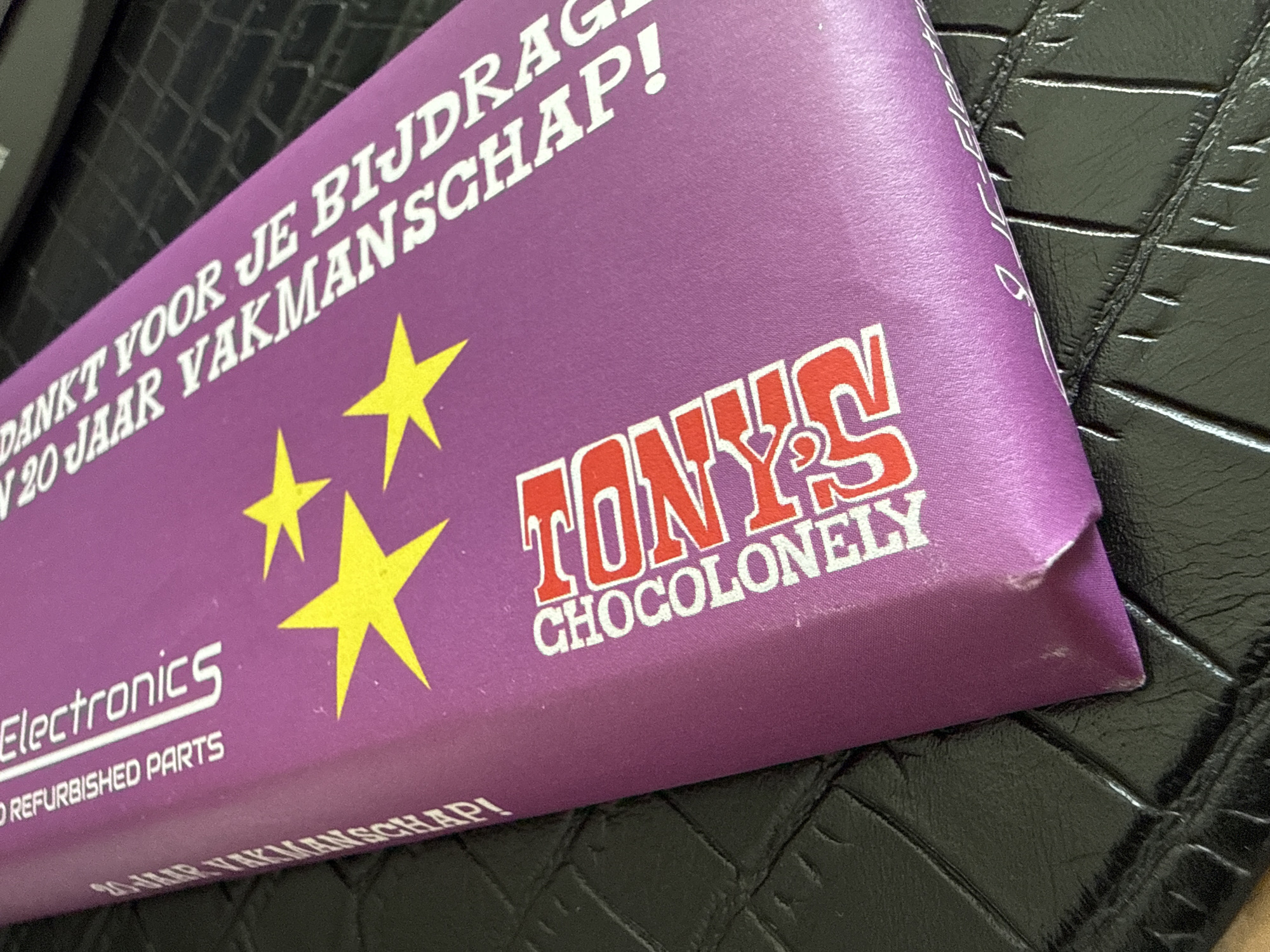 The image shows a close-up, angled view of a purple chocolate bar wrapper from Tony's Chocolonely. The wrapper is lying on a textured black surface, possibly a leather-like material. Text on the wrapper is in white and red. In white, it reads "DANKT VOOR JE BIJDRAGE VAKMANSCHAP!" and "REFURBISHED PARTS" along the left edge. In red, "TONY'S CHOCOLONELY" is prominently displayed in large, stylized letters. Three yellow stars are scattered across the purple background. There are no people visible in the image, and the location in Marum, Netherlands, is not depicted.