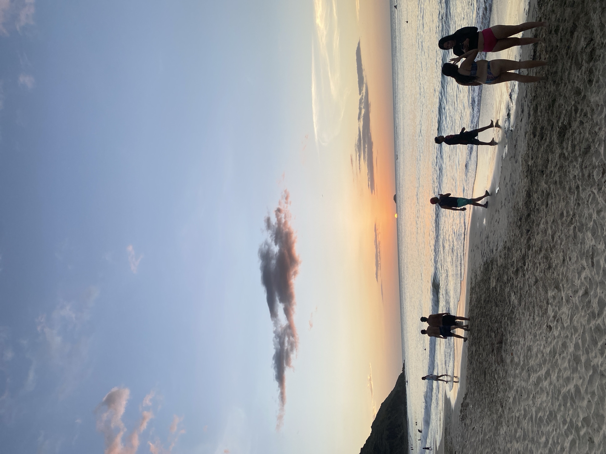 The image captures a serene sunset at Playa El Yaque in Pedro González, Venezuela. The sky is painted with soft hues of pink, orange, and blue, with scattered clouds. The sun is low on the horizon, casting a warm glow over the ocean. Gentle waves lap at the sandy shore, where several people are seen strolling and interacting. In the foreground, a group of four individuals stands on the wet sand, with two women in bikinis facing each other and engaged in conversation, while two men walk away from the camera. Further down the beach, a few other figures are visible, some walking along the water's edge. The coastline is bordered by a lush green hill on the left, suggesting the natural beauty of the area. The overall atmosphere is peaceful and relaxing, indicative of a typical evening at a popular beach destination.