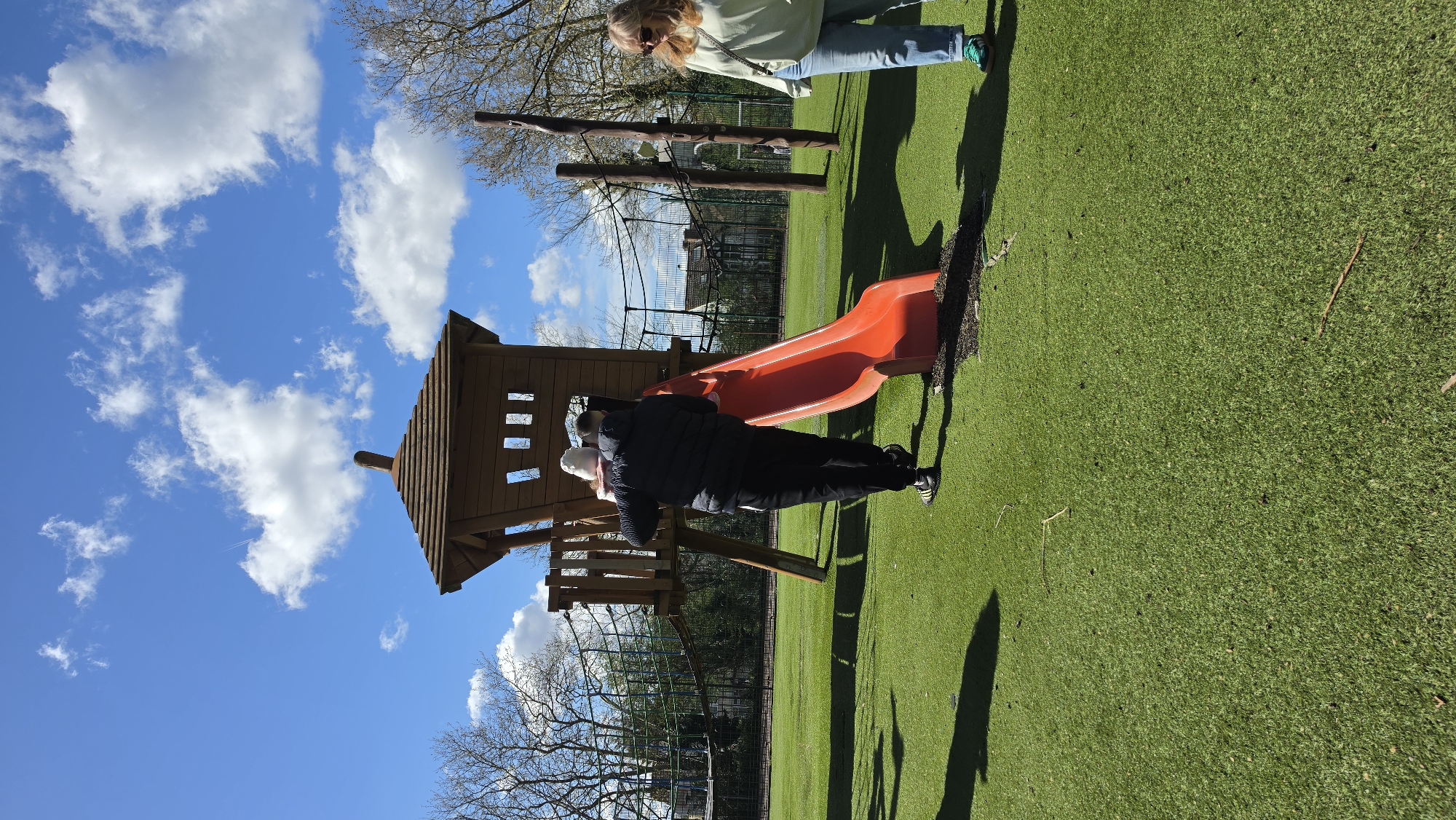 A low-angle shot shows an outdoor playground. A person in a black jacket helps a child in a white hat at the top of an orange slide attached to a wooden playhouse. The playhouse has a small, peaked roof and cutout windows. In the background, a woman wearing light-colored jeans and a light green jacket stands near wooden climbing structures. The playground has artificial green turf. A fence is visible in the mid-ground, with bare trees behind the fence. The sky is blue with white, puffy clouds. Based on the style of the play equipment and fencing, this is in the United Kingdom. Given that there is green grass, this is probably in London.