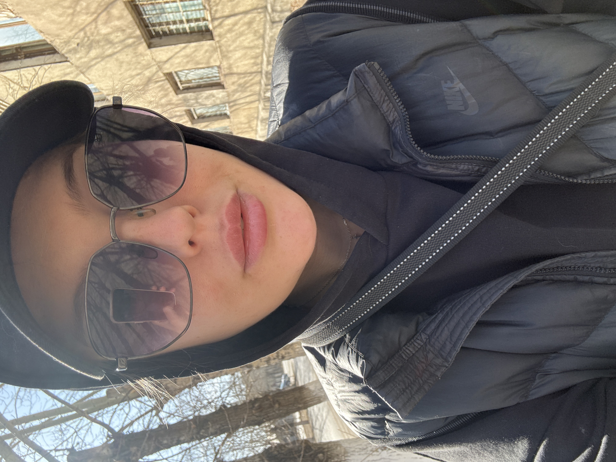 An upward-angled outdoor selfie captures a person in Borzya, Russia, likely during a cool, sunny day in late autumn, winter, or early spring. The image, originally rotated, shows the subject from the shoulders up, looking slightly upwards.
The person wears a dark (likely black) hooded garment beneath a black puffer jacket, on which a faint "NIKE" logo is visible on the left chest area. A black cap is worn, partially visible on the head. Large, squarish sunglasses with dark, reflective lenses obscure most of the face, though full, light-pink lips are visible below them. A black strap with white dotted lines, possibly from a bag or camera, crosses diagonally across the jacket.
In the background, against a bright sky, bare tree branches are prominent on the right side. On the left, a light-colored building with multiple windows can be seen, its surface adorned with distinct shadows of tree branches, indicating strong sunlight. The overall impression is a casual, everyday moment captured outdoors.