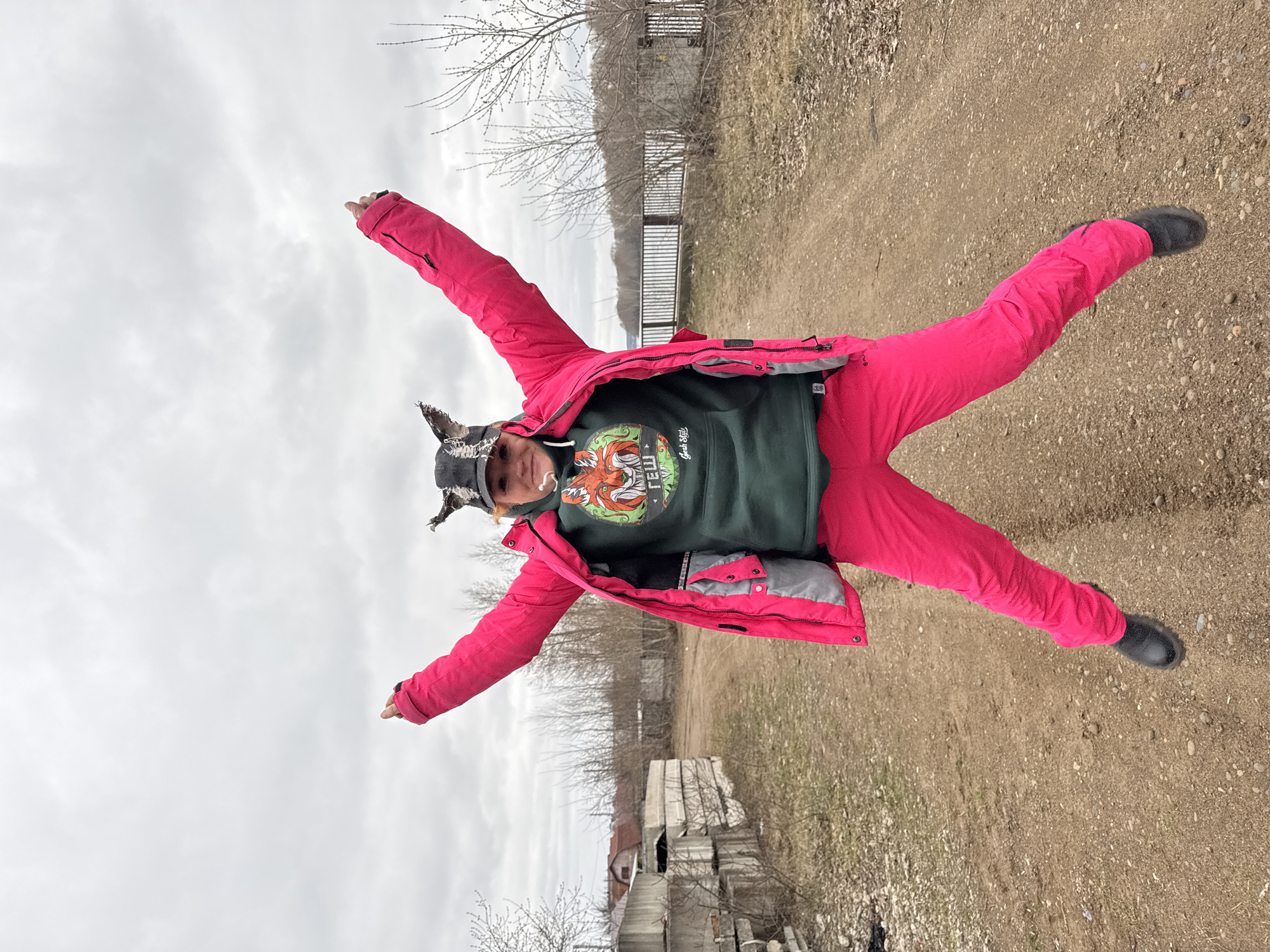 A person in pink outerwear and a hat with ear flaps lies on their back on a dirt path in Chaikovsky, Russia. The person’s arms and legs are extended, creating a star shape. The background includes trees, a fence, and a cloudy sky. Concrete structures are visible in the background.