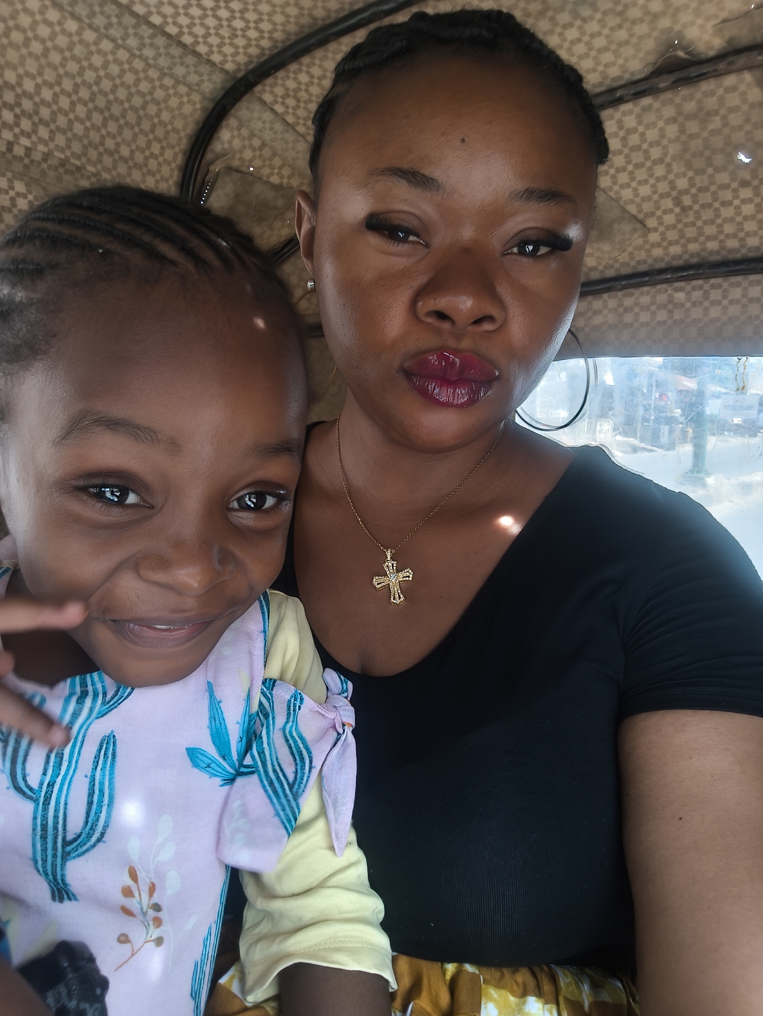 A close-up portrait captures an adult woman and a young child, likely a mother and daughter, seated inside a vehicle. The setting appears to be a form of public transport common in Nigeria, such as an auto-rickshaw (keke Napep), indicated by the distinctive brown and tan checkered fabric ceiling and black metal frame visible above them. Bright daylight suggests it is daytime and sunny outside, with a blurry street scene visible through a window to the right.
The woman, positioned behind the child, has dark skin, braided hair, and is wearing a black top. She sports dark red, glossy lipstick, noticeable false eyelashes, and a golden necklace with a prominent, diamond-studded cross pendant. Her expression is calm and somewhat serious.
The child, in the foreground, also has dark skin and braided hair. They are wearing a light pink and yellow long-sleeved shirt patterned with blue cacti and other botanical elements. The child is looking towards the camera with a wide, joyful smile, eyes bright and full of cheer. Their right hand is partially visible, appearing to point or gesture playfully. The overall mood is one of warmth and happiness, particularly from the child, while the woman maintains a more composed demeanor. There is no visible text in the image.