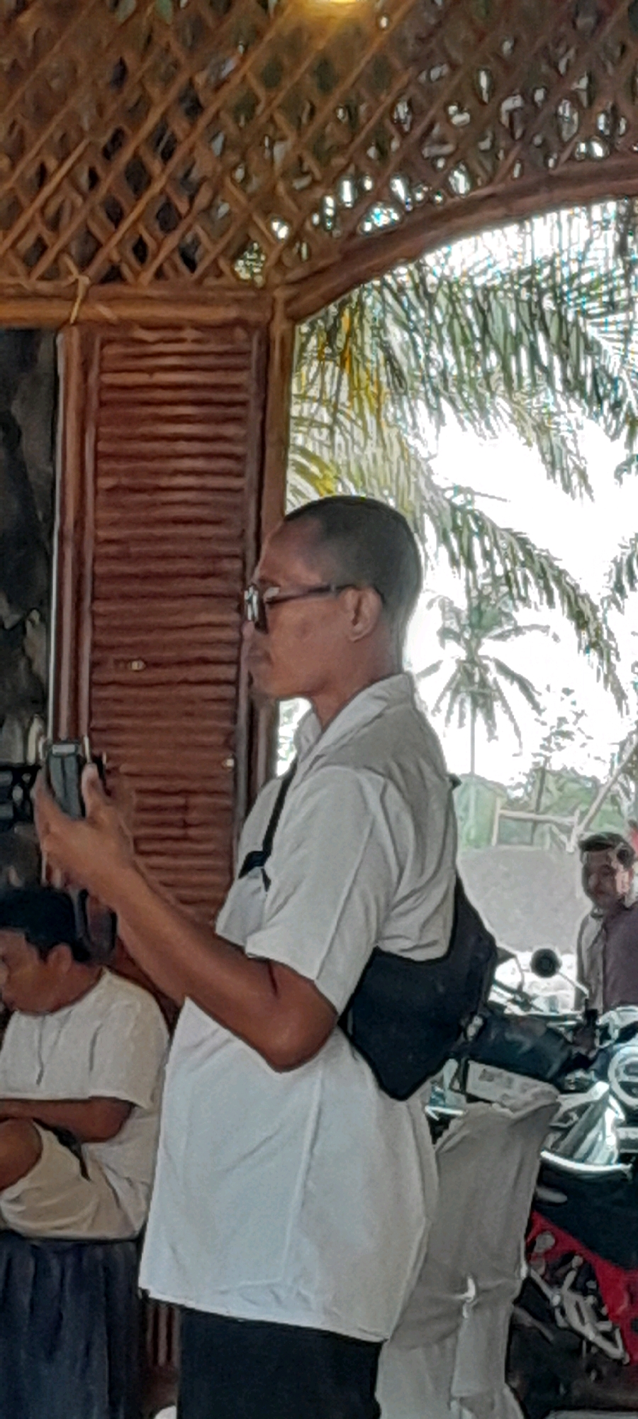 The image shows a man holding a cell phone in his left hand and looking to his left. He is wearing glasses, a white short-sleeved shirt, and a dark-colored sling bag across his chest. He is standing under a bamboo-thatched roof structure with a wooden wall with horizontal slats to his left. In the background, palm trees are visible, suggesting a tropical or subtropical outdoor environment. Another person is partially visible in the background to the right, sitting on a motorcycle. The setting appears to be a casual outdoor gathering or seating area, possibly a cafe or restaurant in Padang, Indonesia, given the presence of palm trees and the architectural style.