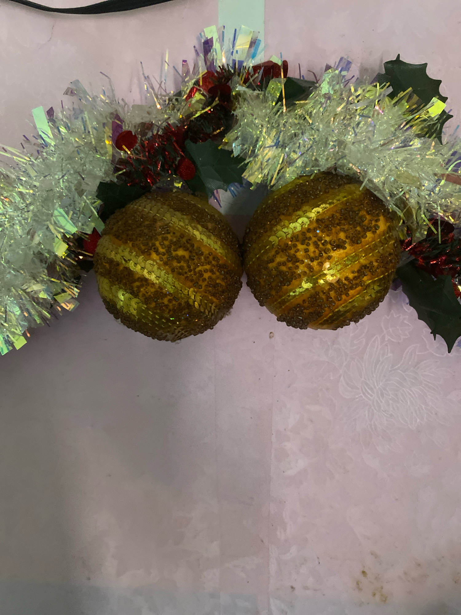 The image depicts a close-up, overhead view of a Christmas garland with two golden, sequined ornaments against a light pink, textured background. The garland features silver tinsel, red berries, and dark green holly leaves. The ornaments are spherical and have a textured surface covered in small, brown sequins arranged in diagonal bands. The background appears to be a fabric or wallpaper with a subtle floral pattern. The location is not specified within the image or its description.