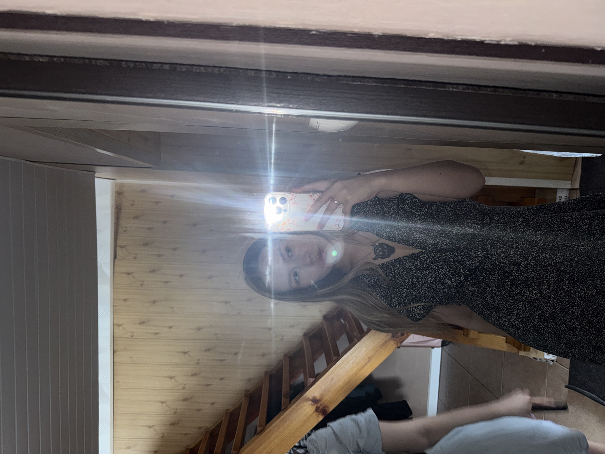 A young woman is taking a mirror selfie in what appears to be a residential hallway or stairwell. She is wearing a dark, patterned dress and a necklace with a pendant. Her left arm, holding a smartphone, is visible, and a bright flash from the phone illuminates the scene. In the background, a wooden staircase with visible steps and a banister is present, along with wood-paneled walls. The overall setting suggests an indoor, possibly domestic environment in Zhigalki, Russia.