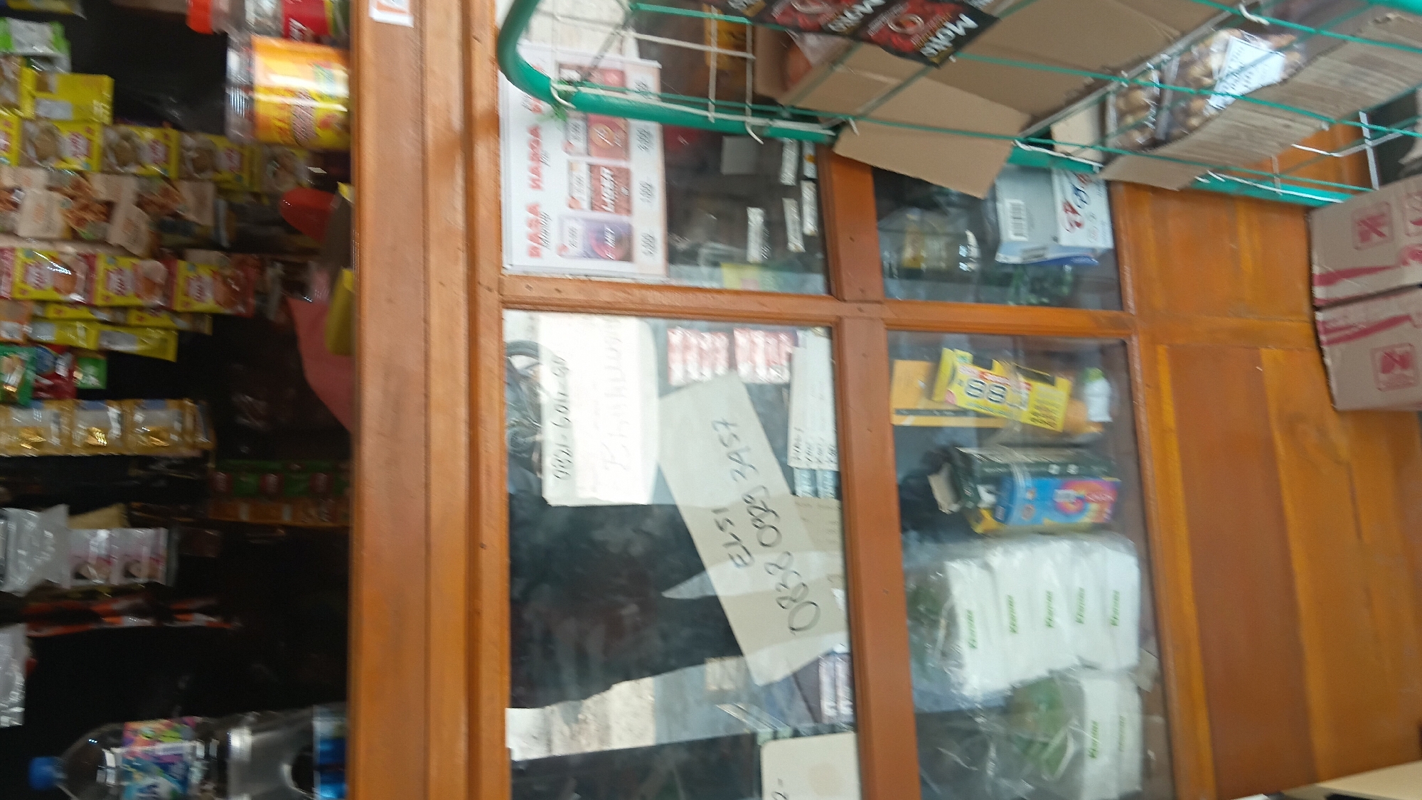 The image shows a dimly lit storefront display case filled with various products. In the foreground, a wooden frame surrounds a glass panel displaying a variety of items. On the left, a partially visible person is reflected in the glass, their dark clothing and legs evident. A white piece of paper with handwritten text, including the numbers "0838 0829 3457" and "ELSI," is affixed to the glass.
Behind this, shelves within the display case hold packaged goods, including what appear to be tissue boxes, cans, and yellow-packaged items with visible text. To the right, another section of the display features a promotional poster with text in Indonesian, including "RASA HARGA" (Taste Price) and product names that are difficult to discern. A green metal rack holding more products, possibly snacks or small items, is also visible on the right.
The overall impression is that of a small convenience store or warung in Prabumulih, Indonesia, displaying a range of everyday consumer goods. The lighting is uneven, with some areas well-lit by an unseen source, while others are in shadow. The reflection of the person suggests they may be the owner or a customer interacting with the display.