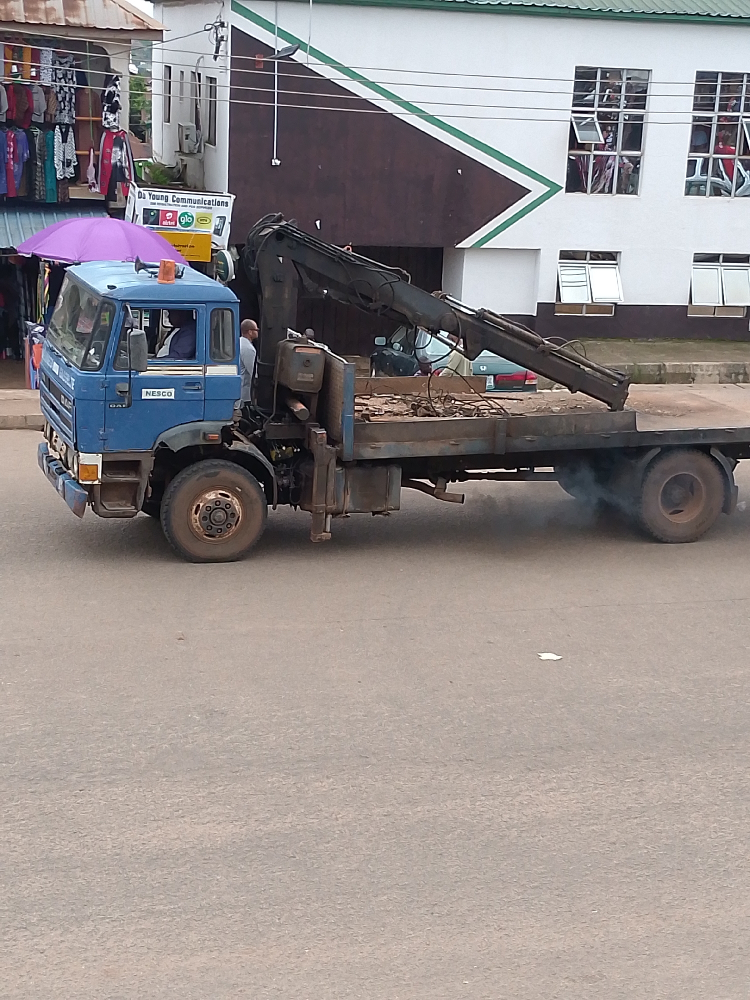 A blue DAF truck with a crane is parked on a paved road in front of a building with a dark brown and white facade. The truck has a flatbed loaded with debris and is emitting a plume of dark exhaust. A purple umbrella is positioned over the driver's side of the truck. A sign above the umbrella reads "Da Young Communications" with company logos. In the background, several people are visible through the windows of the building. The scene appears to be in an urban environment.