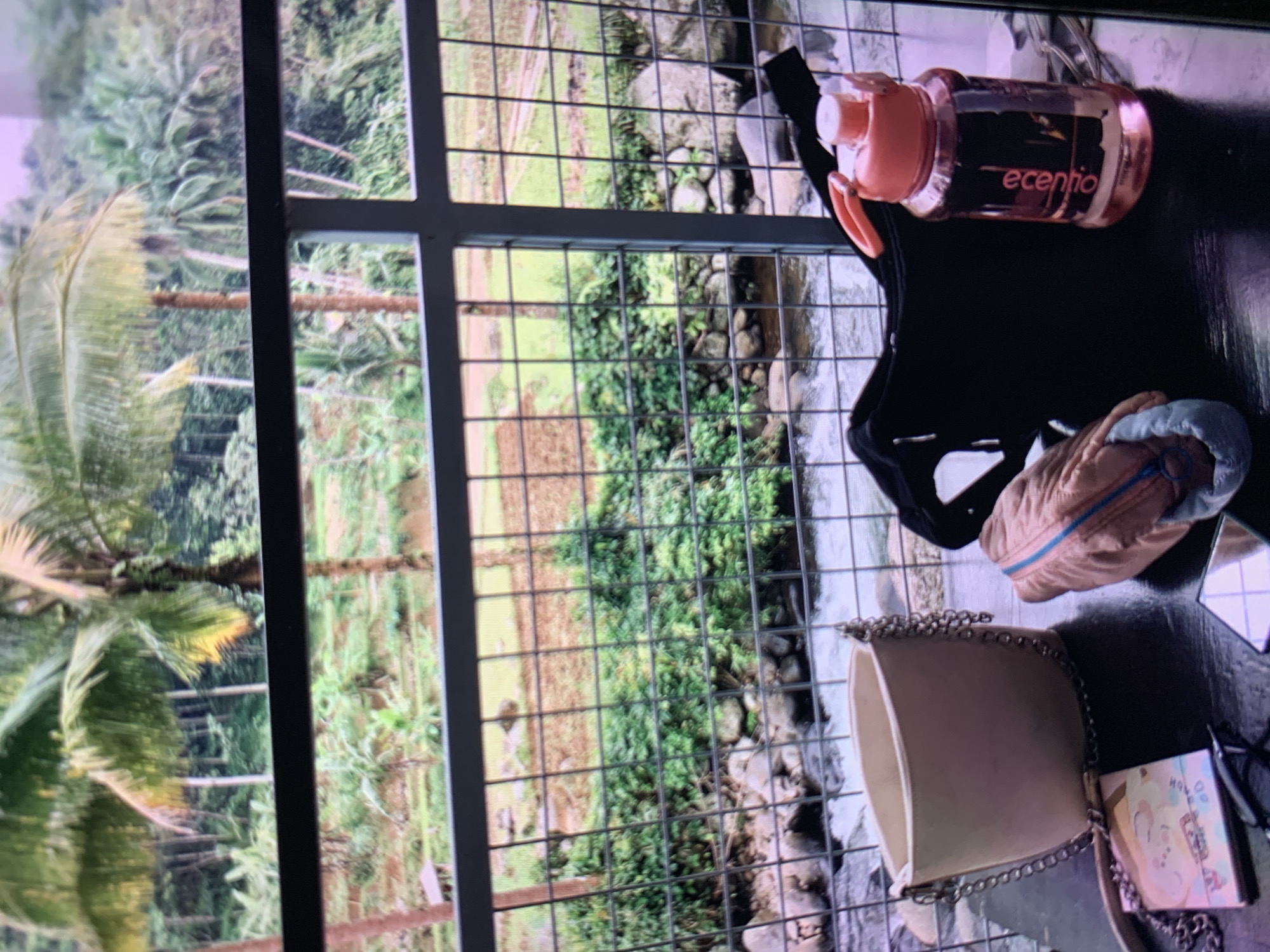 An eye-level shot captures a table on a balcony overlooking a lush green landscape in Ciamis, Indonesia. The scene is dominated by the natural beauty of palm trees and dense foliage, with a river flowing through rocky terrain in the foreground. On the table, a light-colored purse with a silver chain strap rests next to a black fanny pack. A pink water bottle with an orange handle sits beside them. A small notebook and what appears to be a pair of glasses are also visible on the table. The fence in front of the table suggests a viewpoint or a safety barrier. The overall impression is of a tranquil setting, possibly a cafe or a private residence, where one can relax and enjoy the scenic surroundings.