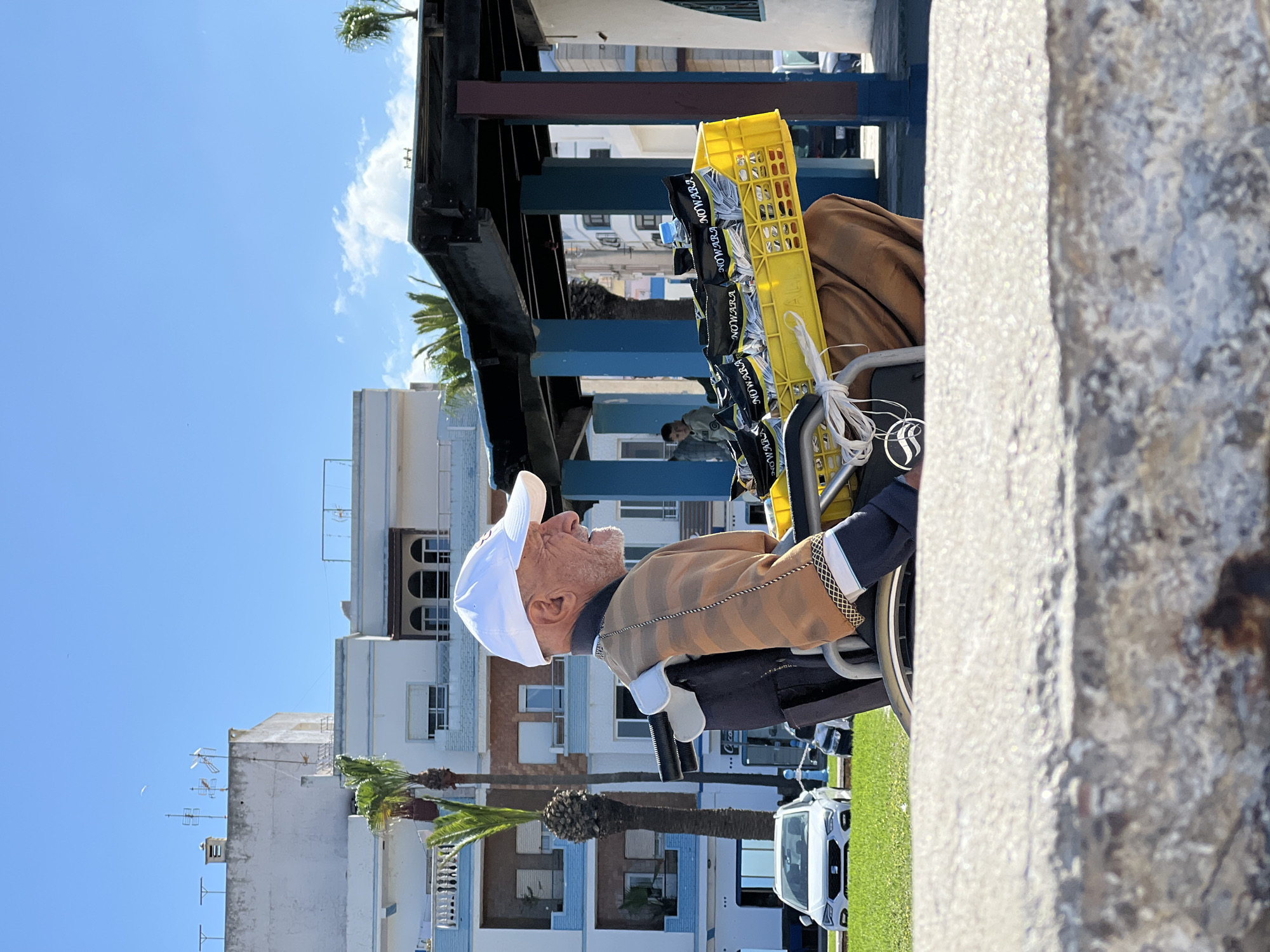 The image depicts an elderly man in a wheelchair, facing left, wearing a white cap and brown striped top. Behind him, a yellow crate filled with individually packaged items, possibly food or goods for sale, is attached to the wheelchair. The setting appears to be an urban area, likely in M'Diq, Morocco, as suggested in the instructions. The backdrop includes buildings with white and blue facades, palm trees, and a covered structure with dark wooden beams. A modern white car is visible in the lower part of the image. The photo is taken from an angle, with a concrete structure visible on the right side, partially obscuring the view. The sky is clear and blue, indicating a sunny day.