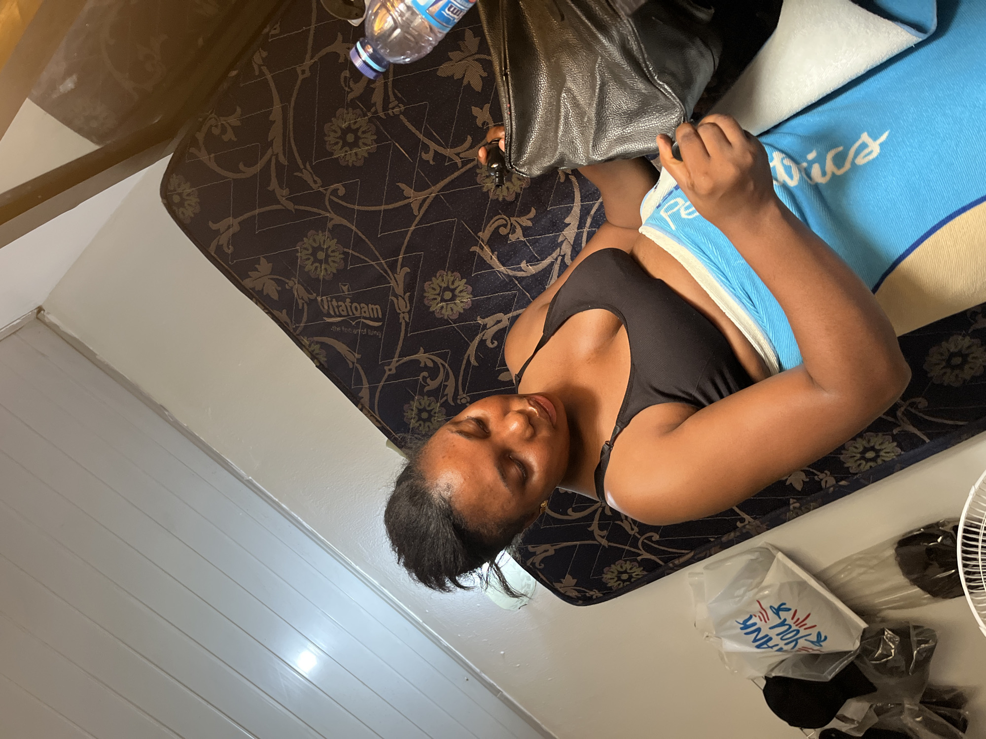 A woman is lying on a Vitafoam mattress with a patterned, dark blue and gold cover, positioned diagonally in a room with white, horizontally-paneled walls. She is wearing a black top and has a light blue blanket with white writing partially covering her body. Next to her on the mattress is a black leather bag and a plastic water bottle. Adjacent to the mattress on the floor, there is a white plastic bag with "THANK YOU" printed on it, alongside dark clothing and a white fan. The image appears to be taken in an indoor setting, potentially a room or apartment, though further location-specific details within Abuja, Nigeria are unavailable from the image.