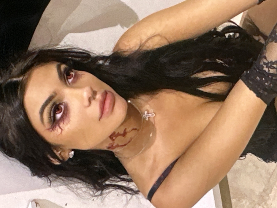 A close-up, chest-up portrait features a young adult female with long, dark, wavy hair, posing in an indoor setting within the city of Novosergievka, Russia. She has dark skin pigmentation. Her eyes are enhanced with red contact lenses, heavy dark eyeliner, long false eyelashes, and red-toned makeup styled to resemble a spiderweb or splattered liquid extending from the outer corners towards her temples. Dark shadowing is present beneath her eyes. Her lips are covered in a light pink, possibly glossy, lipstick. On the right side of her neck, beneath the jawline, red liquid resembling blood is visible in drips and splatters. She wears a thin, clear choker necklace with a silver-colored cross pendant and a small, clear stud earring in her right ear. Her attire includes a dark, possibly black, lacy or textured garment with a thin strap visible over her left shoulder, and a black lace glove or sleeve on her right arm. The background is a plain, light-colored surface, indicative of an interior space, with some dark shadows on the right.