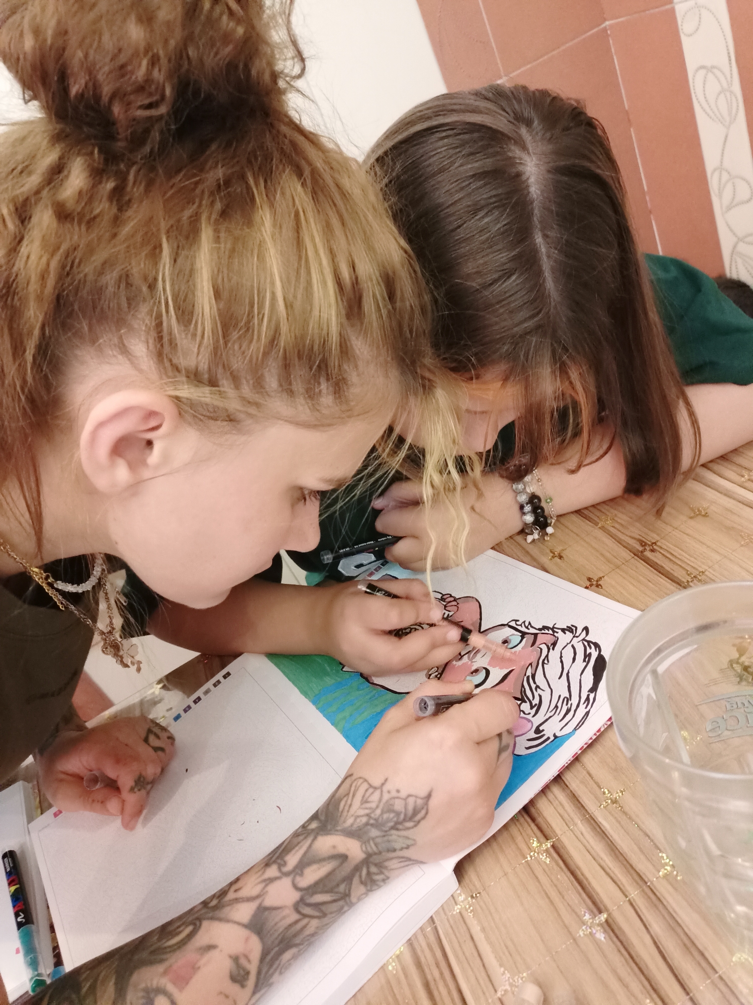 Two young people, one with blonde hair in a bun and the other with dark brown hair, are leaning over a table, coloring a picture in a notebook. The person with blonde hair has tattoos on their arm and hand and is holding a marker, coloring the face of a cartoon character. The person with dark hair is also holding a marker and appears to be coloring the same picture. The table is made of wood with a striped pattern on the wall behind them. There is a glass jar to the right of the notebook. The scene suggests a casual, creative activity taking place indoors. There is no information to suggest this location is in Almaty, Kazakhstan.