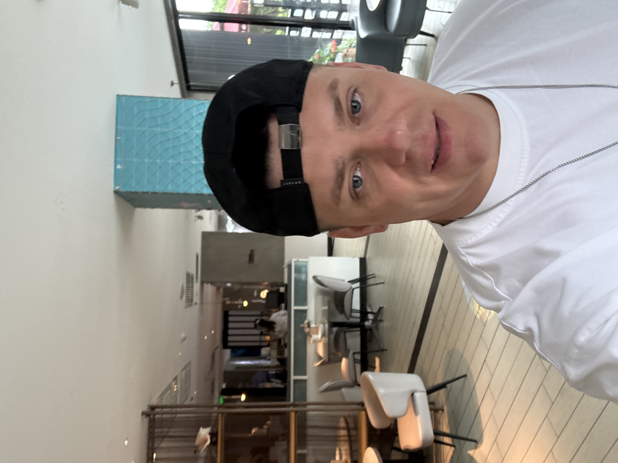 The image is a close-up selfie of a man wearing a black baseball cap and a white t-shirt, taken inside a brightly lit, modern restaurant. The man's face, with blue eyes and light skin, is the primary focus. He is wearing a silver chain around his neck. The background reveals a seating area with tables and chairs, and a counter where a person is visible. The flooring is tiled in a light color with darker lines. A distinctive turquoise tiled column is present in the background. The overall ambiance suggests a contemporary dining establishment. The image was captured in Qinhuangdao, China.