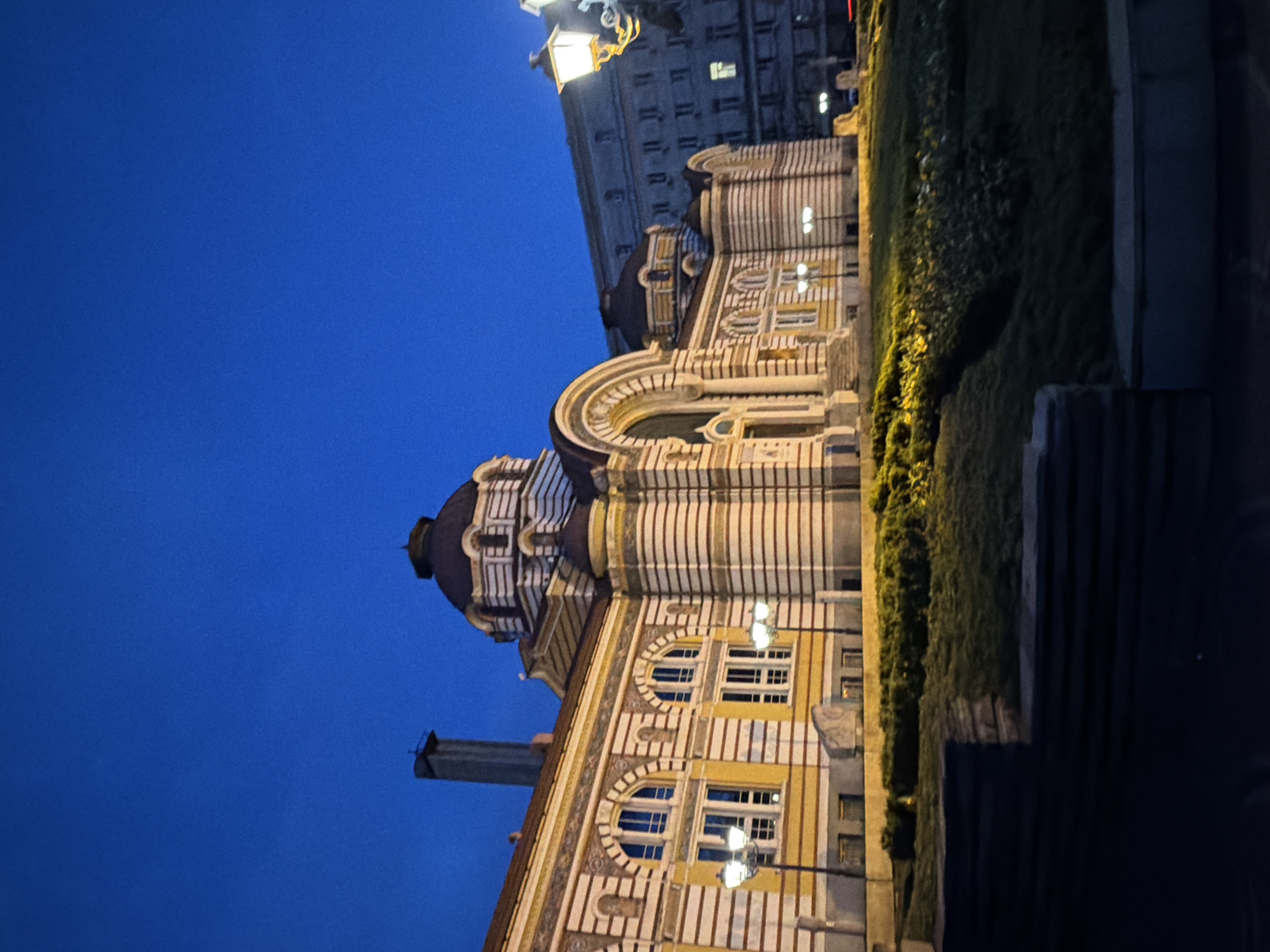 The image captures the Sveti Sedmochislenitsi Church in Sofia, Bulgaria at night. The church is illuminated, showcasing its unique striped architectural style. A building is visible in the background to the right of the top of the Church. The sky is a deep blue. Streetlights are present near the church. A line of shrubbery lines part of the building to the right.