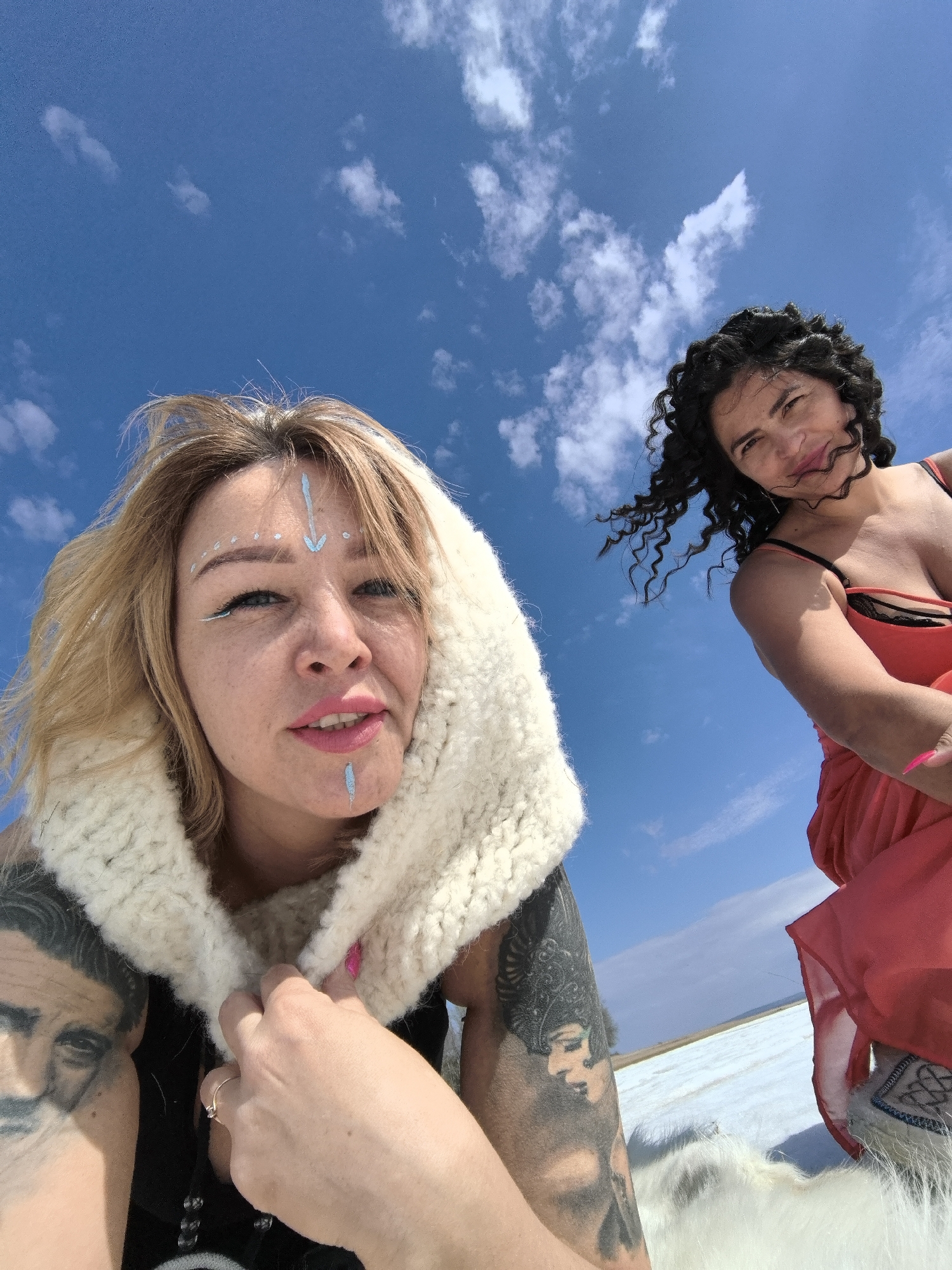 Two women are captured in a low-angle outdoor shot against a bright blue sky dotted with scattered white clouds. The ground beneath them is a vast white expanse, possibly snow or a salt flat, stretching into the distance under the clear daylight in what appears to be Noviy, Russia.
The woman on the left, positioned closer to the camera, has blonde hair and fair skin. She is wearing a thick, cream-colored, fluffy hooded garment. Her face features light blue or white paint, including an arrow pointing down on her forehead, several dots around her eyes, and a vertical line below her lower lip. Her lips are painted bright pink. She has prominent tattoos on both arms; one depicts a man's face, and the other shows a woman with an elaborate headdress. A ring is visible on her right ring finger.
The woman on the right, slightly further back and to the side, has dark, curly hair that appears to be gently blown by the wind. She wears a red top or dress with thin black straps, under which a black lace bra is visible. She offers a subtle smile towards the camera. Her right hand shows bright pink nail polish, and a fur-lined boot with intricate embroidery is visible on her left foot. Both women are looking down towards the camera, suggesting they are posing for a photograph. They appear to be seated or reclined on a white fur or fabric spread on the ground. The weather is sunny and bright, indicative of midday.