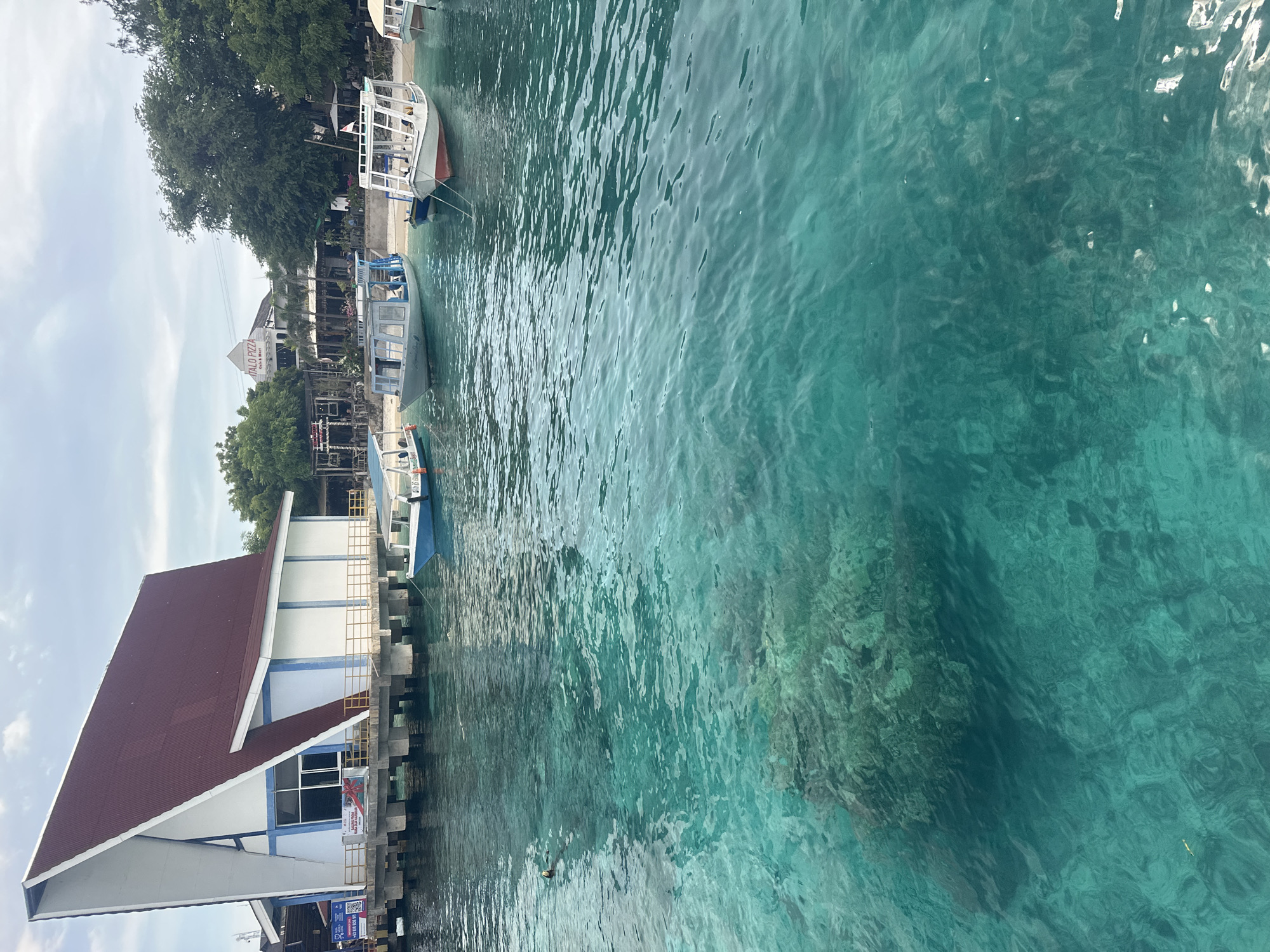 The image depicts a waterfront scene, possibly a harbor or pier in Indonesia. The water is a clear, turquoise color, suggesting shallow depth and good visibility. There are several boats docked alongside a pier or platform extending from a building with a brown roof and white walls. The building appears to be situated directly on the water. The structure is built on a foundation of piers that extend into the water. The image appears to be taken during the day, with soft, diffused sunlight. The location is likely in the city of Unknown, Indonesia.