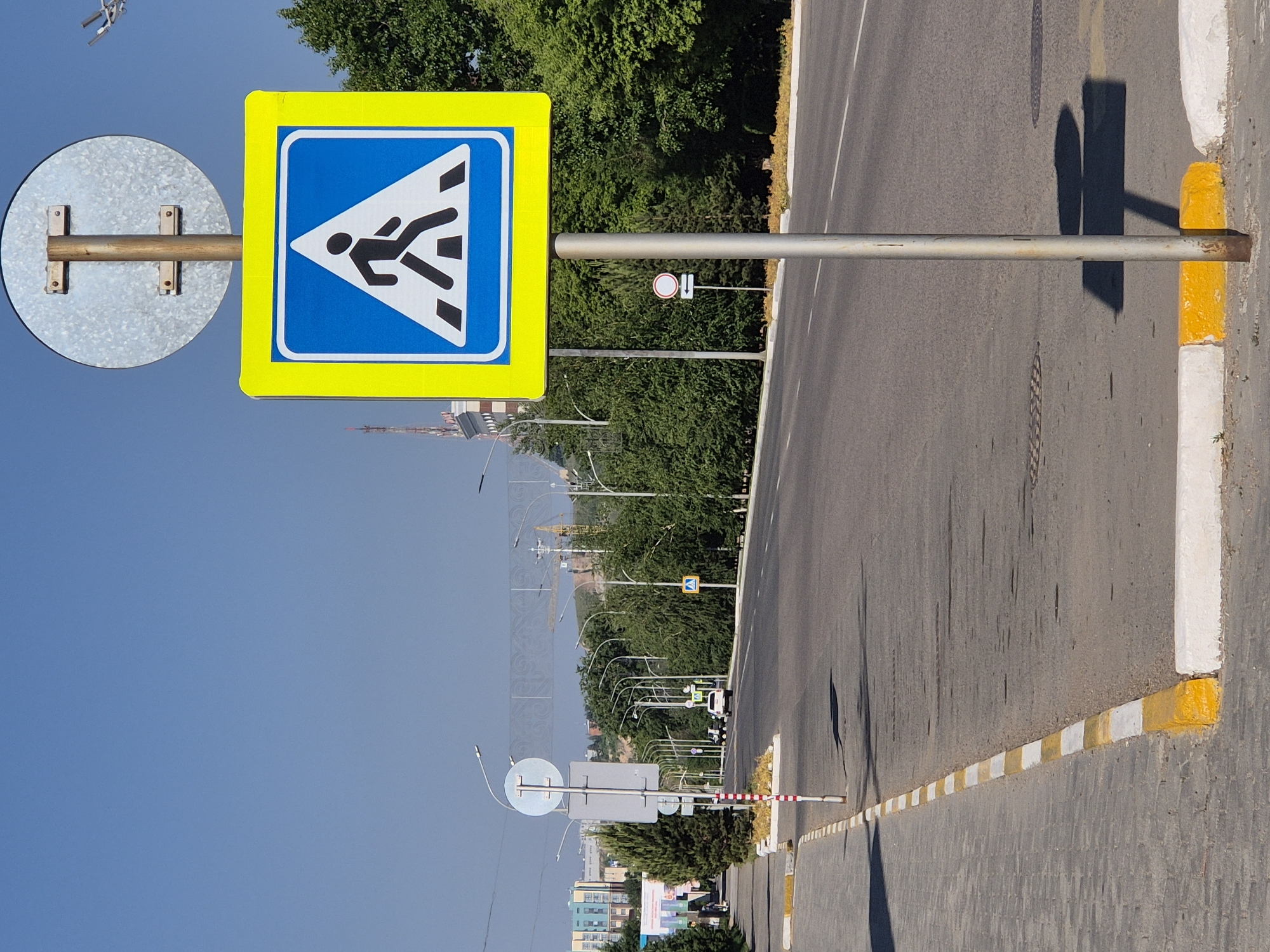 The image depicts a street scene in Kokshetau, Kazakhstan, featuring a pedestrian crossing sign. The sign, rectangular with a yellow border and a blue interior containing a white triangle with a black figure walking across striped bars, is attached to a metal pole. The pole also holds a round metal disc of an indeterminate traffic sign. The street is asphalt, marked with a yellow and white curb. In the background, other traffic signs and streetlights line the road, with trees and buildings visible further away. The sky is clear and blue.