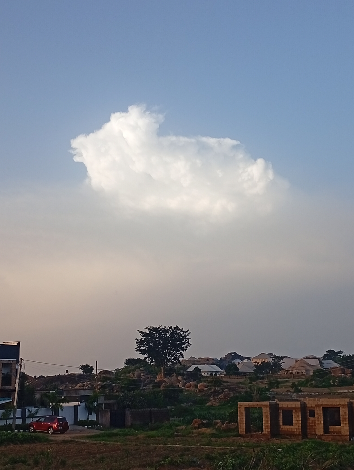 The image depicts a scenic view of Gura Top, Nigeria, under a bright blue sky dotted with a large, fluffy white cloud. In the foreground, a red car is parked near a building under construction, with a single tree standing tall on a rocky hill in the background. The landscape transitions into a cluster of houses with distinctive thatched roofs, nestled amongst the rocky terrain. The overall impression is one of a peaceful, developing community in a rural setting.