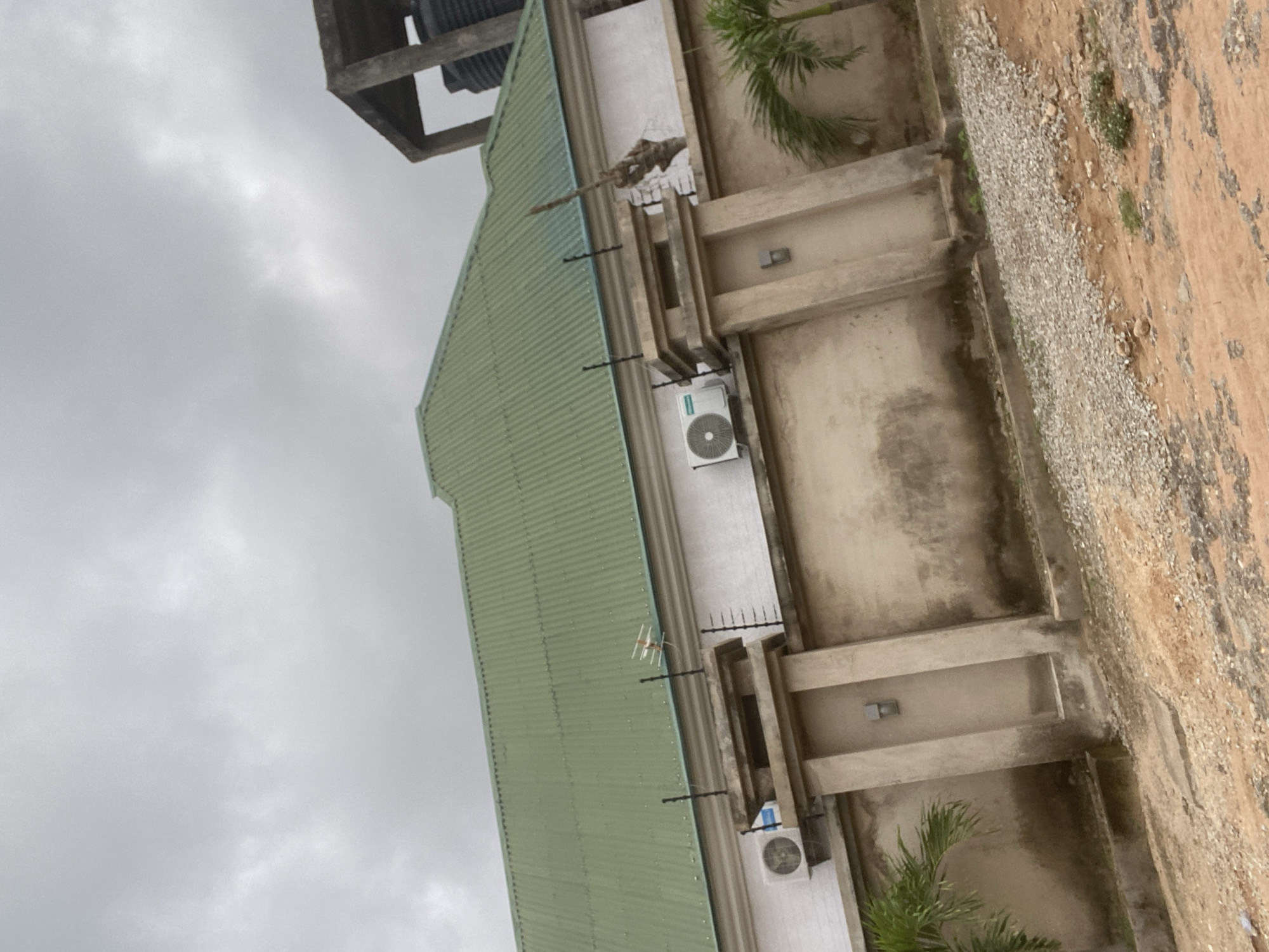 This image depicts a house with a green corrugated metal roof, set behind a concrete wall. The wall has multiple upright concrete pillars and appears to be topped with electric fencing. Two outdoor air conditioning units are visible mounted on the exterior wall of the house. A large black water tank sits atop a concrete structure in the upper right corner of the image. The ground in front of the wall is covered with gravel and dirt. The sky is overcast with gray clouds. There are no people or animals visible in the image. The scene is located in Gbaza, Nigeria.