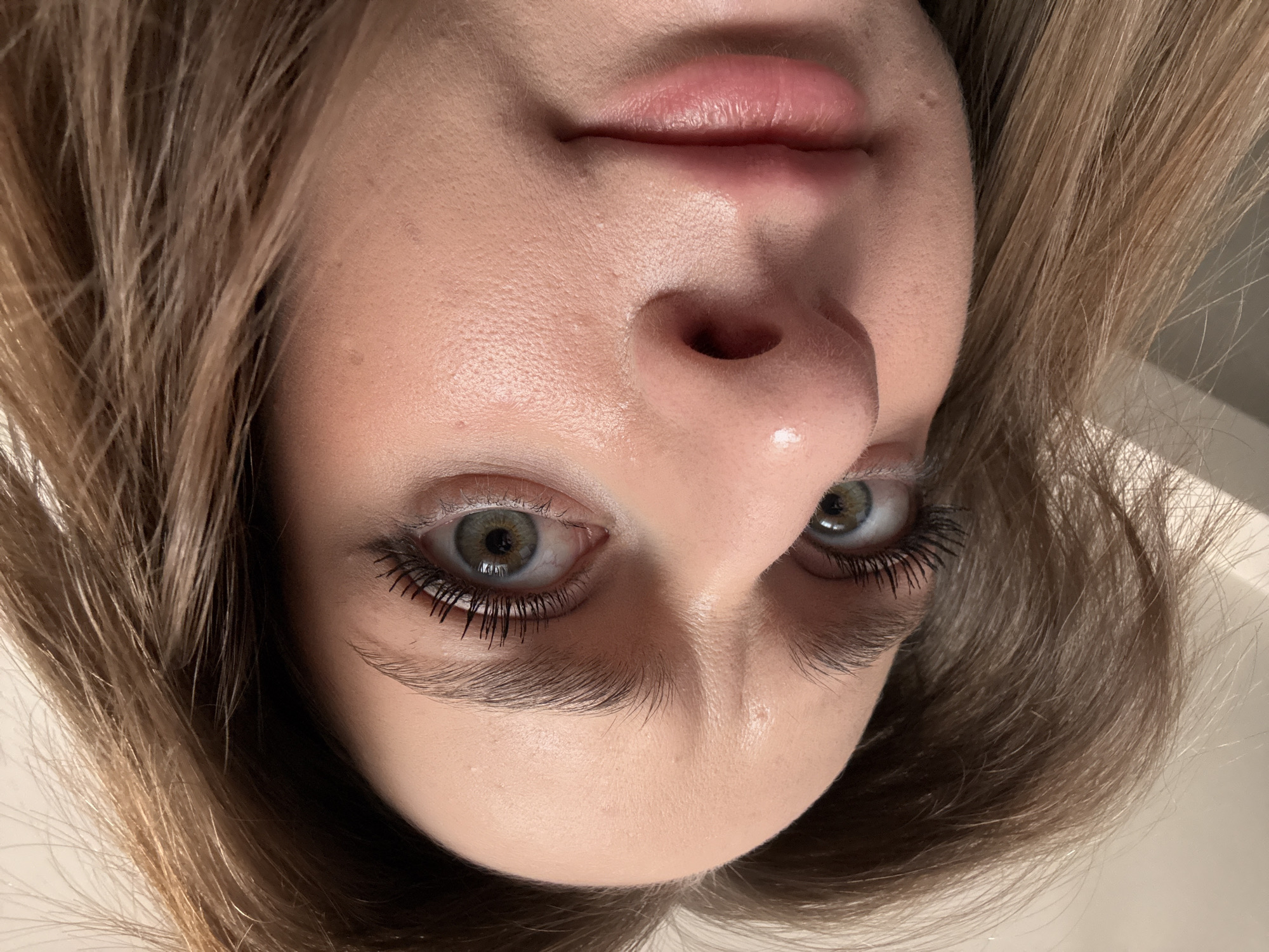 Close-up, inverted view of a woman's face. She has fair skin, light brown hair, and green eyes. She is wearing mascara. The background is a white surface. The location is not evident from the image.