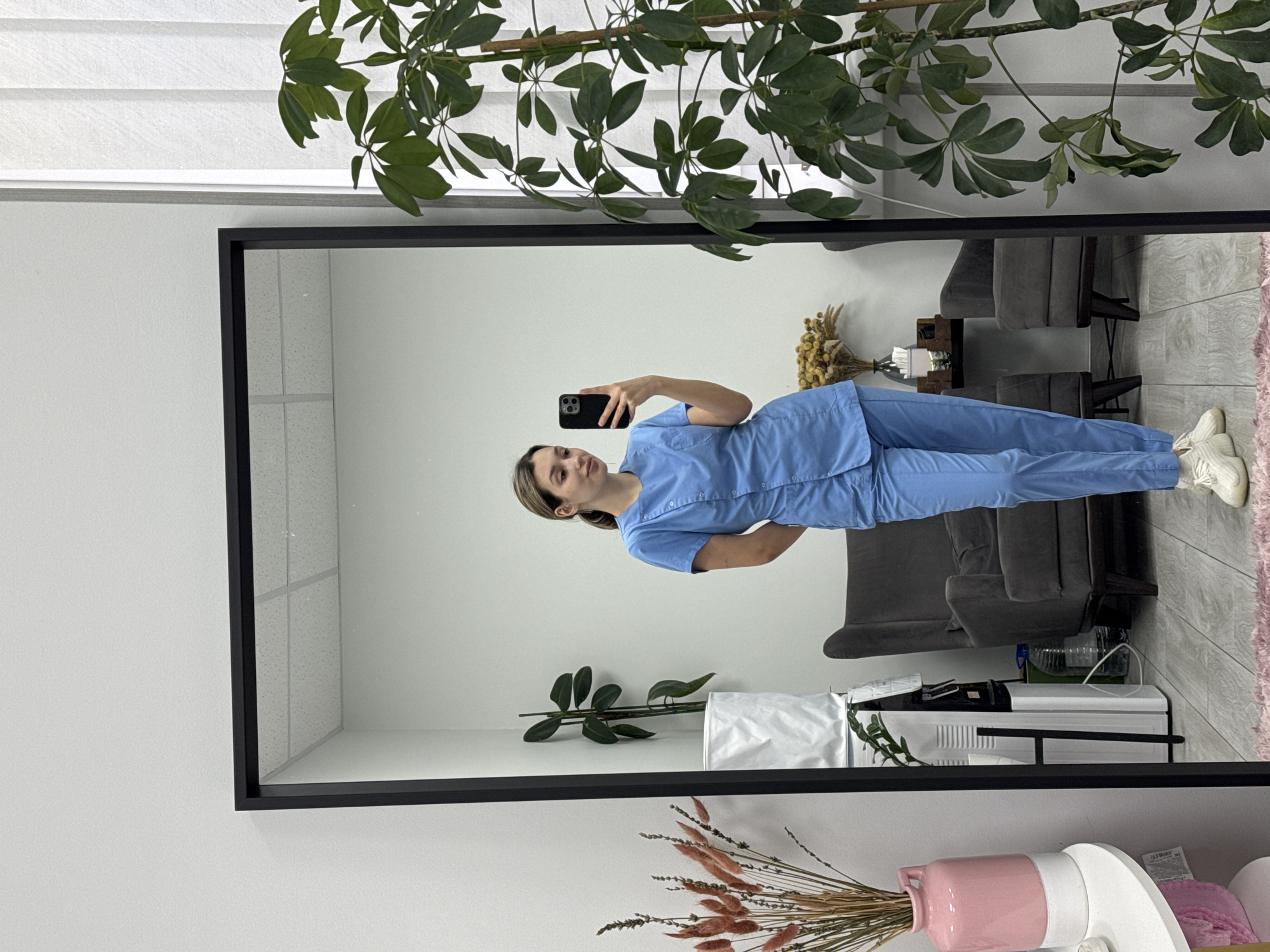 A young woman in light blue scrubs takes a full-body mirror selfie indoors. She is positioned slightly to the left in the large, black-framed mirror, holding a black smartphone to capture her reflection. Her light brown hair is pulled back, and she has a calm, neutral expression. She is wearing white sneakers.
The reflection in the mirror reveals a clean, professional-looking room. Behind her, two gray armchairs or small sofas are arranged, with a dark wooden side table between them. On the table, a dried floral arrangement in a brown vase is visible, along with a white object that appears to be a lamp. The floor in the reflection is tiled, and a light pink rug is partially visible on the right. A large green leafy plant is situated above and to the left of the mirror, with its reflection also appearing inside the mirror, hanging downwards. The ceiling in the reflection features white grid tiles. To the right of the armchairs, a white appliance, possibly an air purifier or medical device, is visible on the floor.
Outside the mirror's frame, the wall is plain white. In the bottom foreground, a pink ceramic vase filled with dried grasses and flowers is visible, along with other blurry objects suggesting a decorative or functional display. The overall setting appears to be a professional office or clinic waiting area, indicated by the scrubs and furniture.