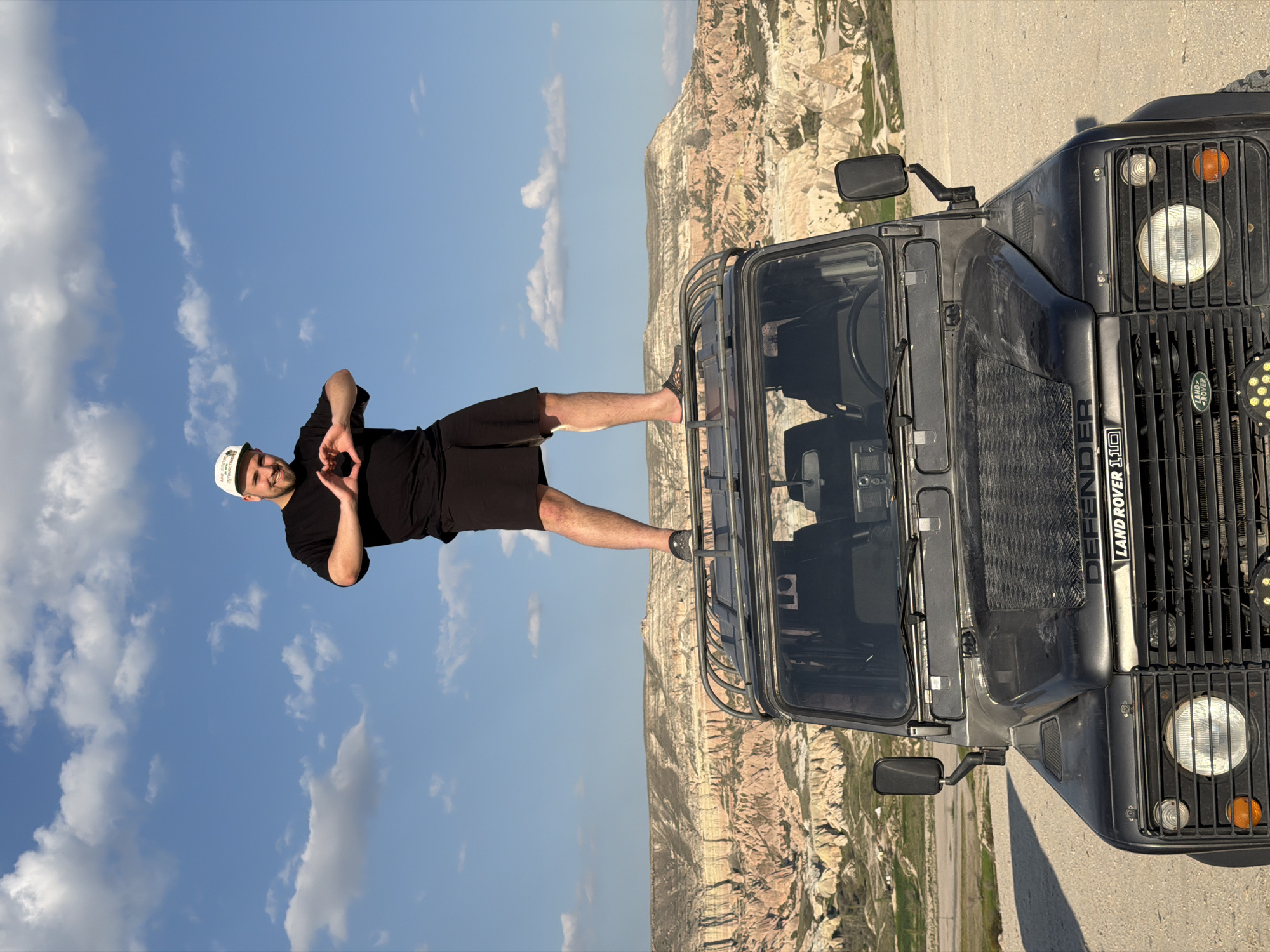 A man is pictured posing dynamically on the side of a dark-colored Land Rover Defender 110, set against a backdrop of unique geological formations under a clear blue sky. The man, likely in his 20s or 30s, has a beard and a smiling expression. He wears a black t-shirt, black shorts, a white cap, and sneakers. He stands with one foot on the vehicle's side step and the other lifted, and his hands are clasped together to form a heart shape, indicating a cheerful or appreciative gesture.
The vehicle is a rugged, dark grey or black Land Rover Defender 110, visible from its front and passenger side. It features a prominent front grille, a large circular headlight on the passenger side, an amber turn signal, and an aftermarket multi-LED auxiliary light. A sturdy roof rack is also visible over the cab. Visible text on the vehicle includes "LAND ROVER DEFENDER 110" on the side of the bonnet/fender and the classic "Land Rover" logo on the front grille.
The background showcases the distinctive, light-colored, eroded rock formations and valleys characteristic of Göreme, Cappadocia, Turkey. The terrain appears dry and dusty. The sky above is a vibrant blue, dotted with scattered white cumulus clouds, suggesting a sunny day. The overall scene conveys a sense of adventure and highlights the striking natural beauty of the location.