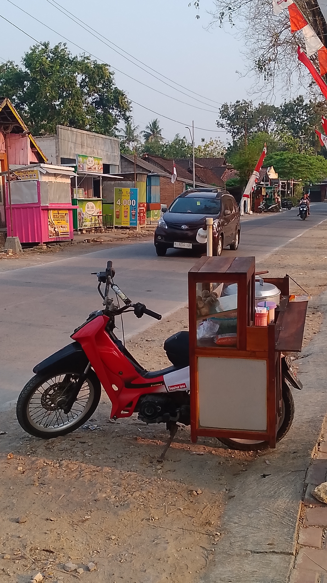In the city of Karanglangu, Indonesia, a red motorcycle with a food cart attached is parked on the side of a paved road. The food cart is constructed of wood and glass and contains various food items, including what appear to be steamed buns in a glass display. The motorcycle itself is a small, sporty model. In the background, there are several small food stalls and buildings, with trees and foliage. Indonesian flags are visible, decorating some of the stalls. A dark-colored SUV is driving on the road, and a person on a motorcycle is also visible in the distance. The scene appears to be set during the late afternoon, with warm lighting casting shadows. The road is bordered by a dirt shoulder and a concrete walkway.