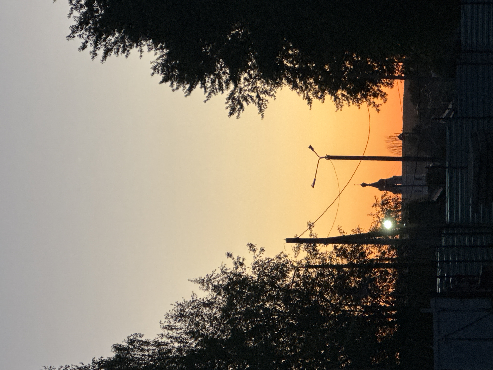 Captured at dusk or dawn in Berezovka, Russia, the photograph features a silhouette of the Russian Orthodox Church against a vibrant orange sky. The view is partially framed by trees with full foliage. Power lines and a streetlight intersect the sky, adding to the urban landscape. A metal fence is visible on the right side of the frame. The light suggests a street view, with the church possibly serving as a local landmark.