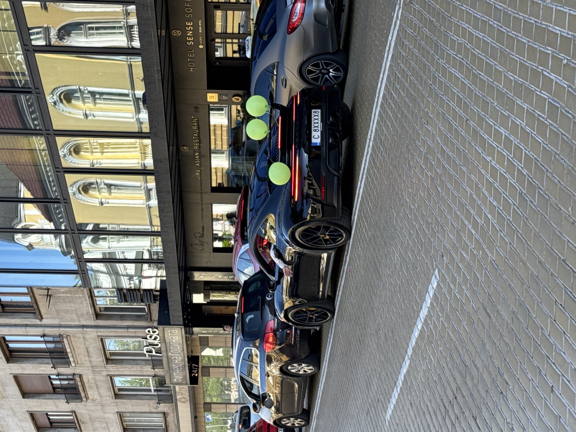 The image shows a city street scene in Sofia, Bulgaria, with several cars parked alongside a building that houses both the Hotel Sense Sofia and the Jiru Asian Restaurant. A black Porsche at the front has three green balloons attached to its back. The Porsche's license plate reads "C 8XXXX8." Next to the restaurant is a fitness center called "Pulse." The buildings reflect the surrounding environment in their windows. The street is paved with patterned brick.