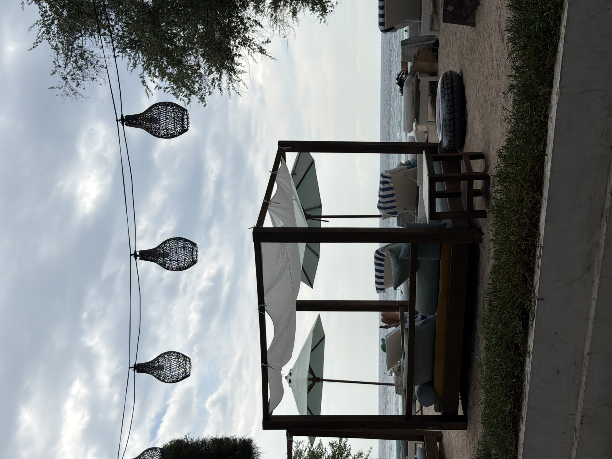 In the city of Unknown, Indonesia, a beach scene unfolds under a cloudy sky. Three decorative wicker pendant lights hang suspended from a wire, casting a rustic ambiance over the sandy expanse. In the foreground, a covered cabana with white fabric drapes over a wooden frame, providing shade. Inside, cushioned seating arrangements are visible, including a long daybed with blue and yellow cushions. Two light blue umbrellas, one of which is partially unfurled, stand near the cabana. Beach chairs with striped blue and white cushions are scattered around the area, some with items on them. The ocean stretches out in the background, with gentle waves lapping at the shore. A section of green foliage lines the bottom of the image, separated by a low concrete barrier. The overall atmosphere suggests a tranquil resort setting.