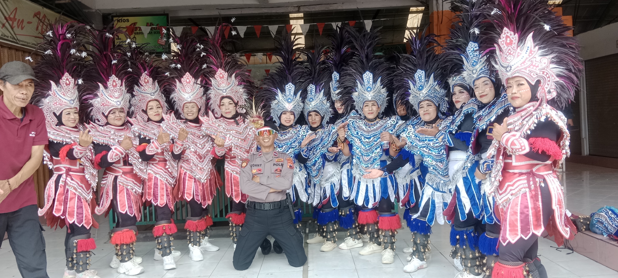 In Magelang, Indonesia, a group of performers in elaborate feathered headdresses and costumes are posing for a photo. The performers are predominantly women, dressed in matching vibrant outfits, some in red and silver, others in blue and silver, adorned with intricate beadwork and fabric. They wear tall, feathered headdresses that frame their faces, and many have gold bangles on their arms. One woman on the far left is wearing a red polo shirt and a dark baseball cap, while a police officer in uniform kneels in the center of the group, arms crossed, wearing sunglasses. The background suggests an outdoor setting, possibly in front of a building with signage, and flags are strung across the top of the scene. The ground appears to be paved.