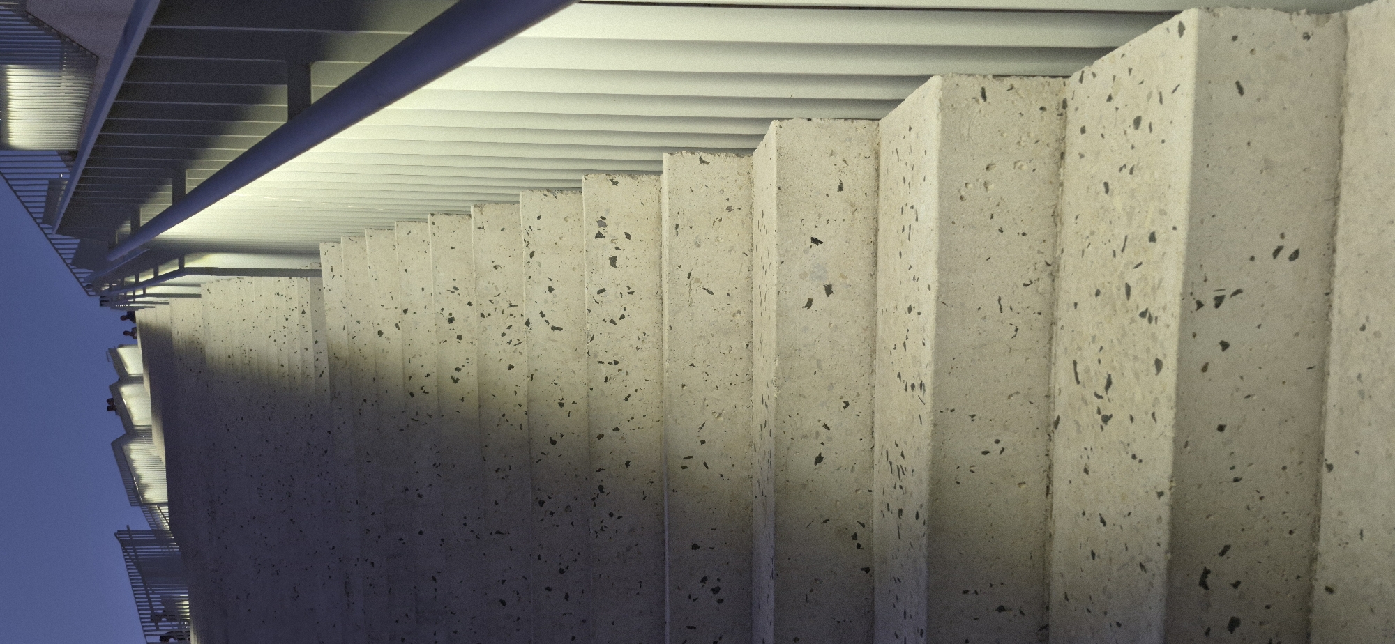 This image shows a set of outdoor concrete stairs illuminated by artificial lighting. The stairs are textured with small, dark aggregate embedded in the concrete. A silver-colored handrail runs along the right side of the stairs, supported by vertical metallic poles. In the background, the sky is a deep blue, indicating dusk or dawn. There are several figures visible on the upper part of the stairs, appearing as silhouettes against the lighter background, suggesting they are walking or standing. The architectural style, with its clean lines and integrated lighting, is characteristic of modern public spaces. While the image itself does not provide explicit information about its location, the request specifies Tirana, Albania. This suggests the stairs are part of a public infrastructure or a modern architectural development within Tirana.