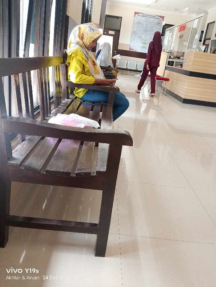 A woman in a yellow jacket and patterned hijab sits on a wooden bench, looking at her phone. Beside her sits another woman in a white hijab. In the background, a woman in a maroon outfit stands near a counter. A large poster hangs on the wall behind them. The scene appears to be a waiting area in a clinic or public building, featuring shiny tiled floors.